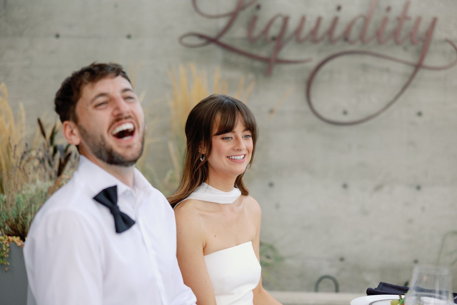 Happy bride and groom laughing together at Liquidity winery, bride wearing halter neck gown and groom in bow tie.