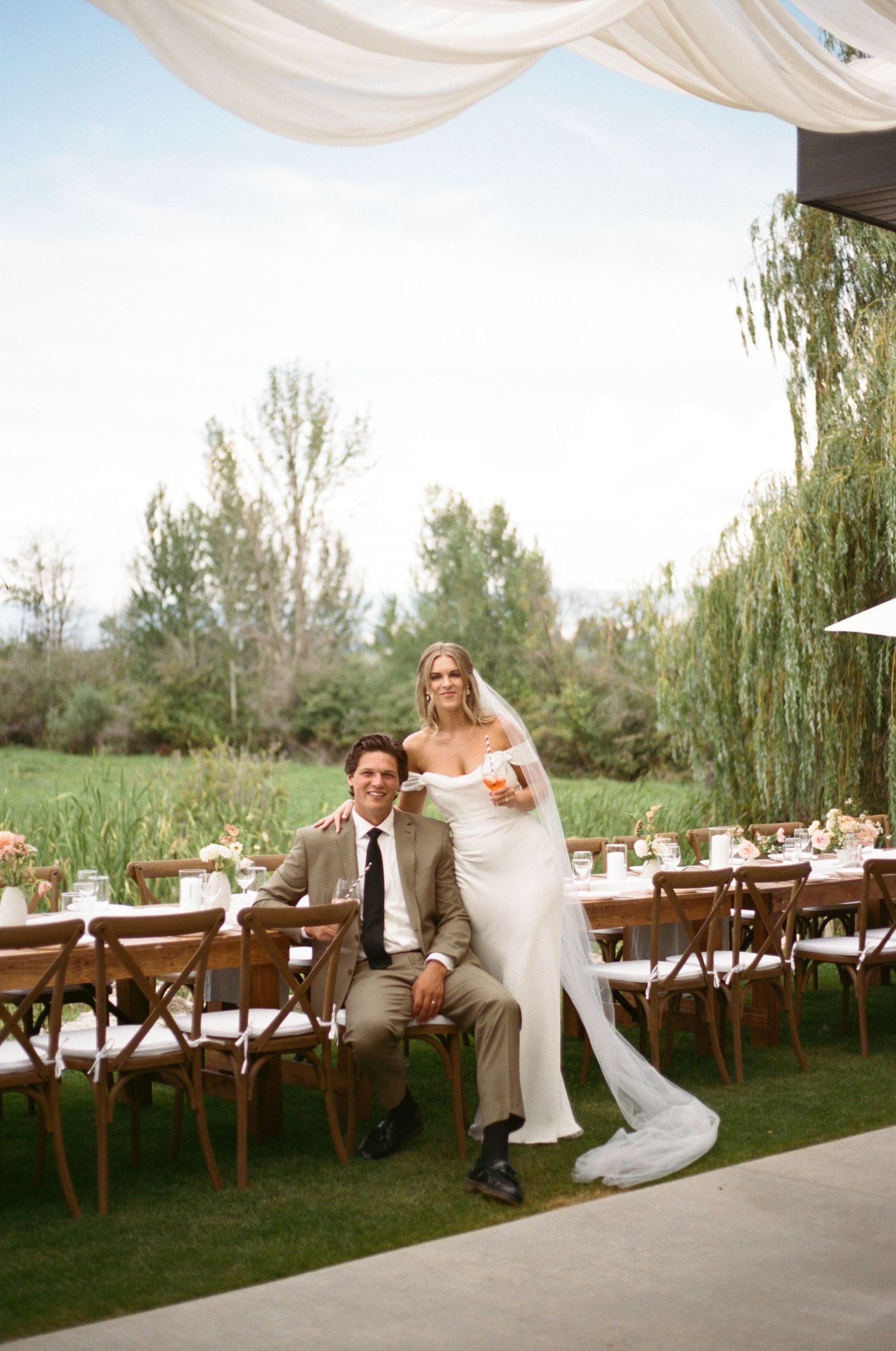 Bride and groom posing at elegant outdoor wedding reception with long wooden tables under a draped canopy.