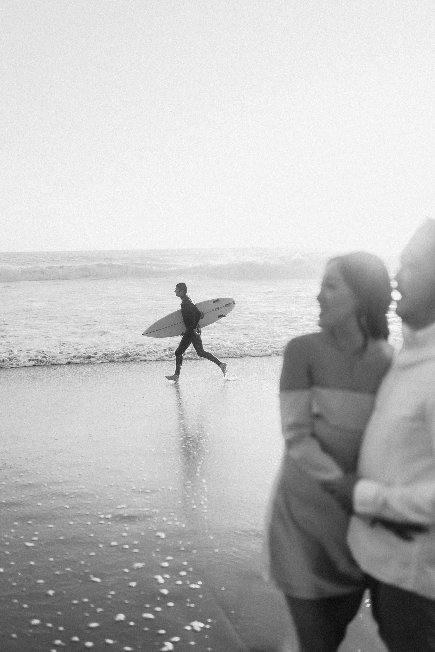 A black and white beach photo of a surfer carrying a board near the shore while a couple stands in the foreground.
