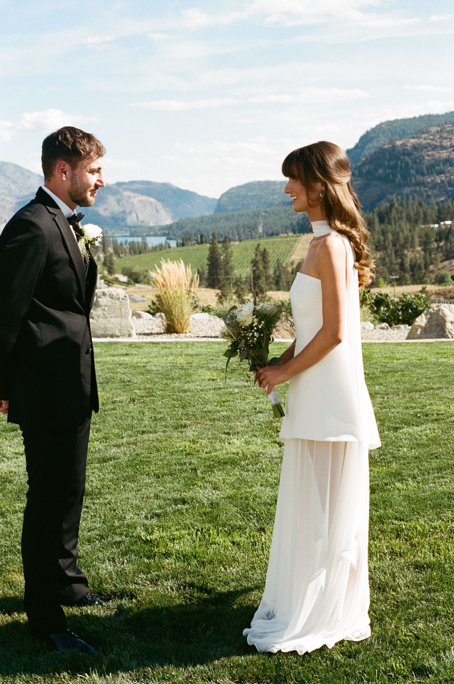 Bride and groom share a joyful moment on green lawn with stunning mountain lake backdrop.