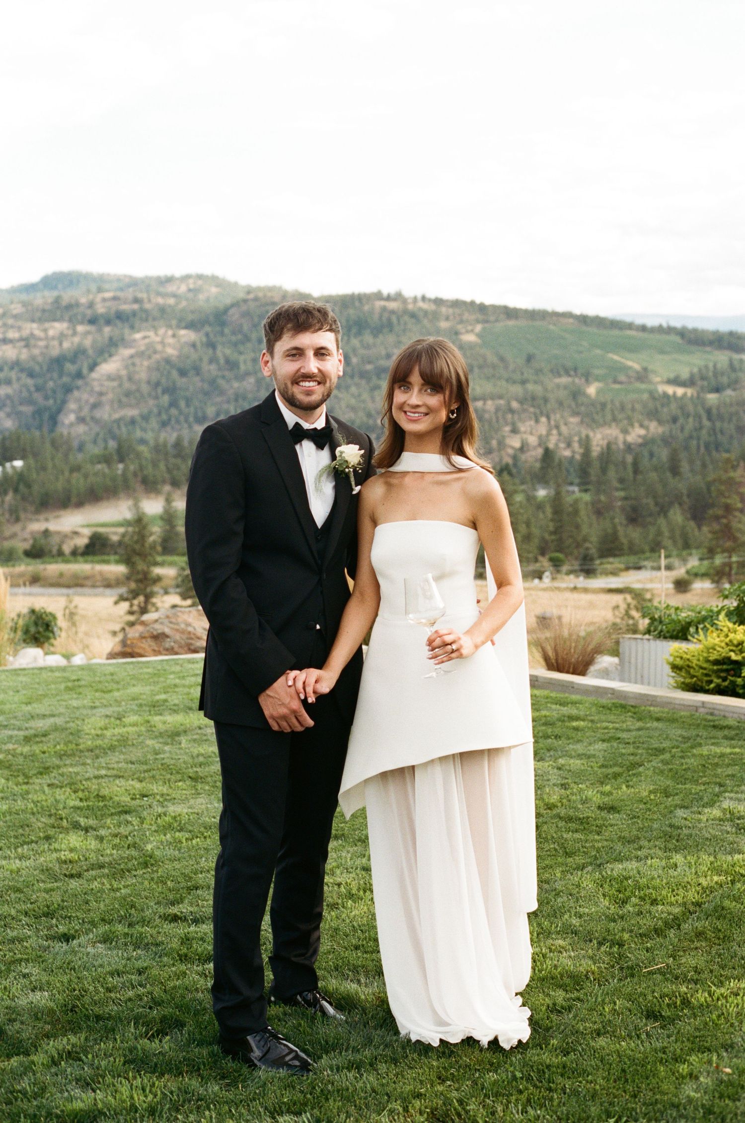 Bride and groom smiling together on green lawn with mountain and forest backdrop at outdoor wedding.