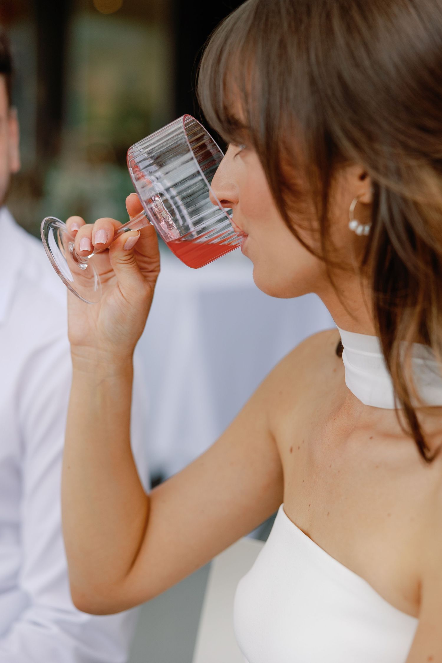 Close-up of bride in white halter gown sipping rose wine from a crystal glass at outdoor wedding reception.
