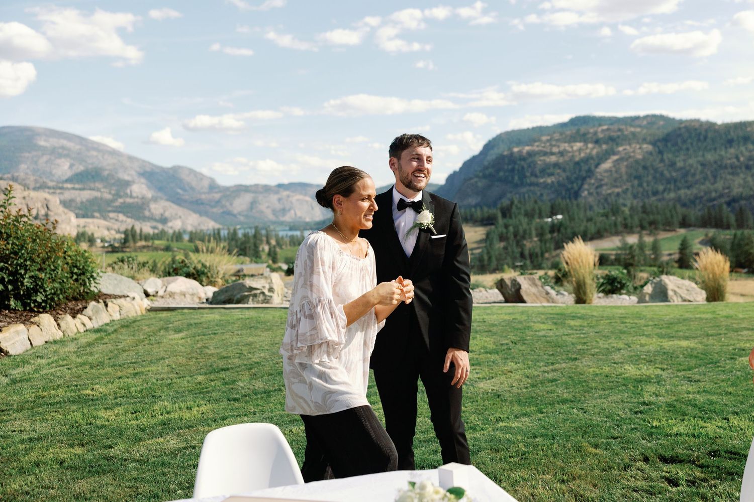 Bride in lace top and groom in tuxedo holding hands at outdoor wedding with mountains.