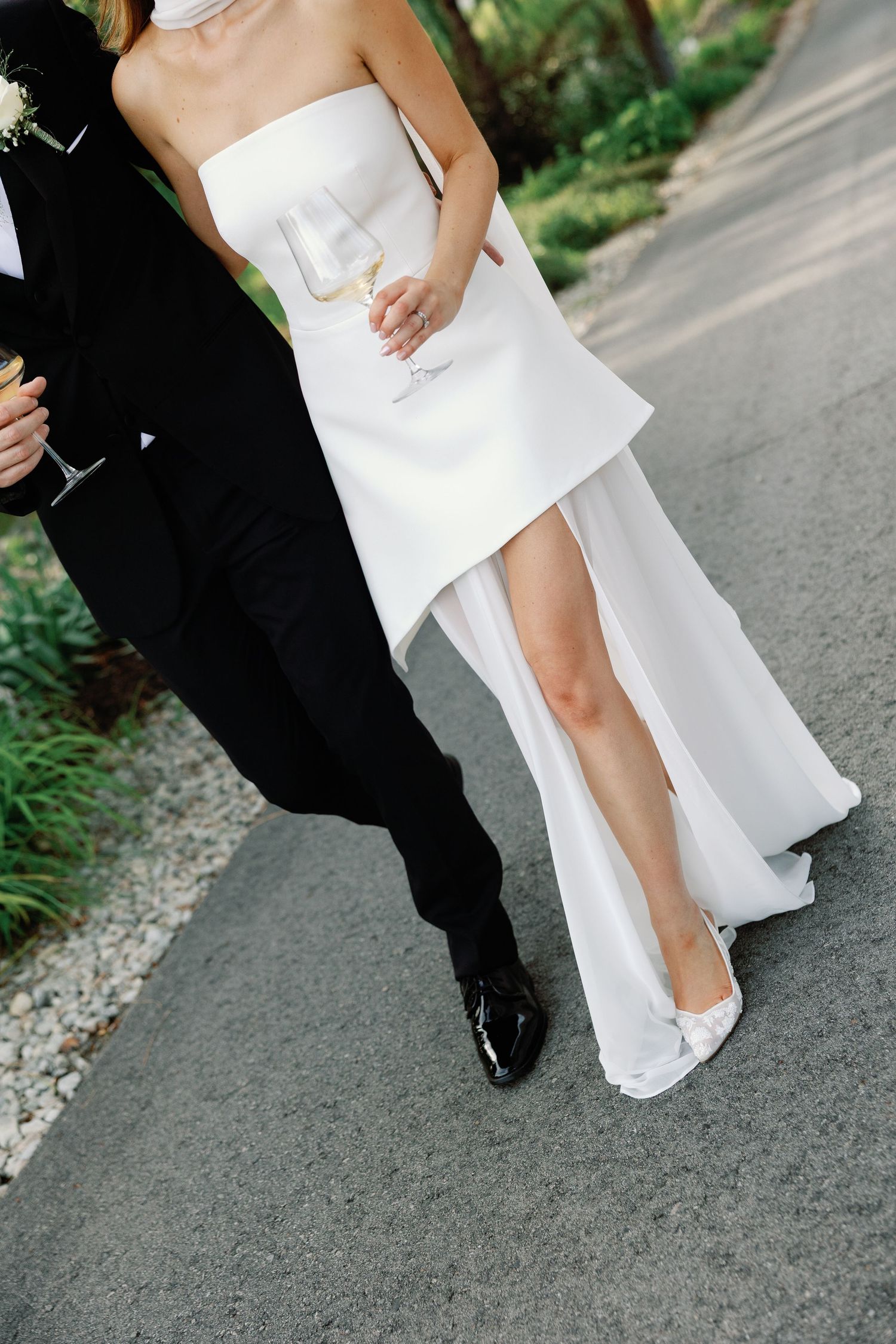 Bride in a strapless white high-low gown walks with groom holding champagne on a garden pathway.