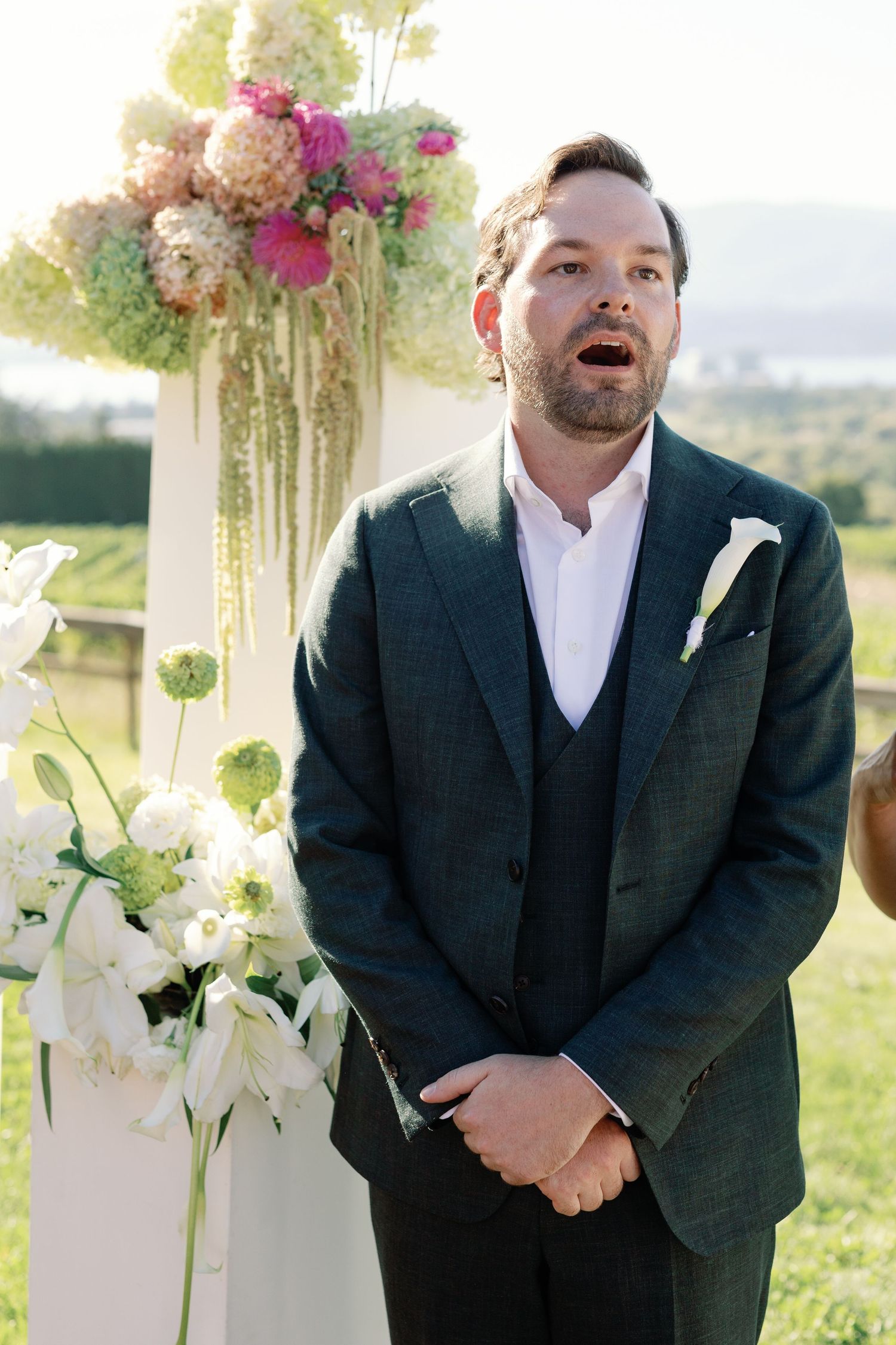 Groom in dark green suit reacting emotionally at the altar during outdoor wedding ceremony.