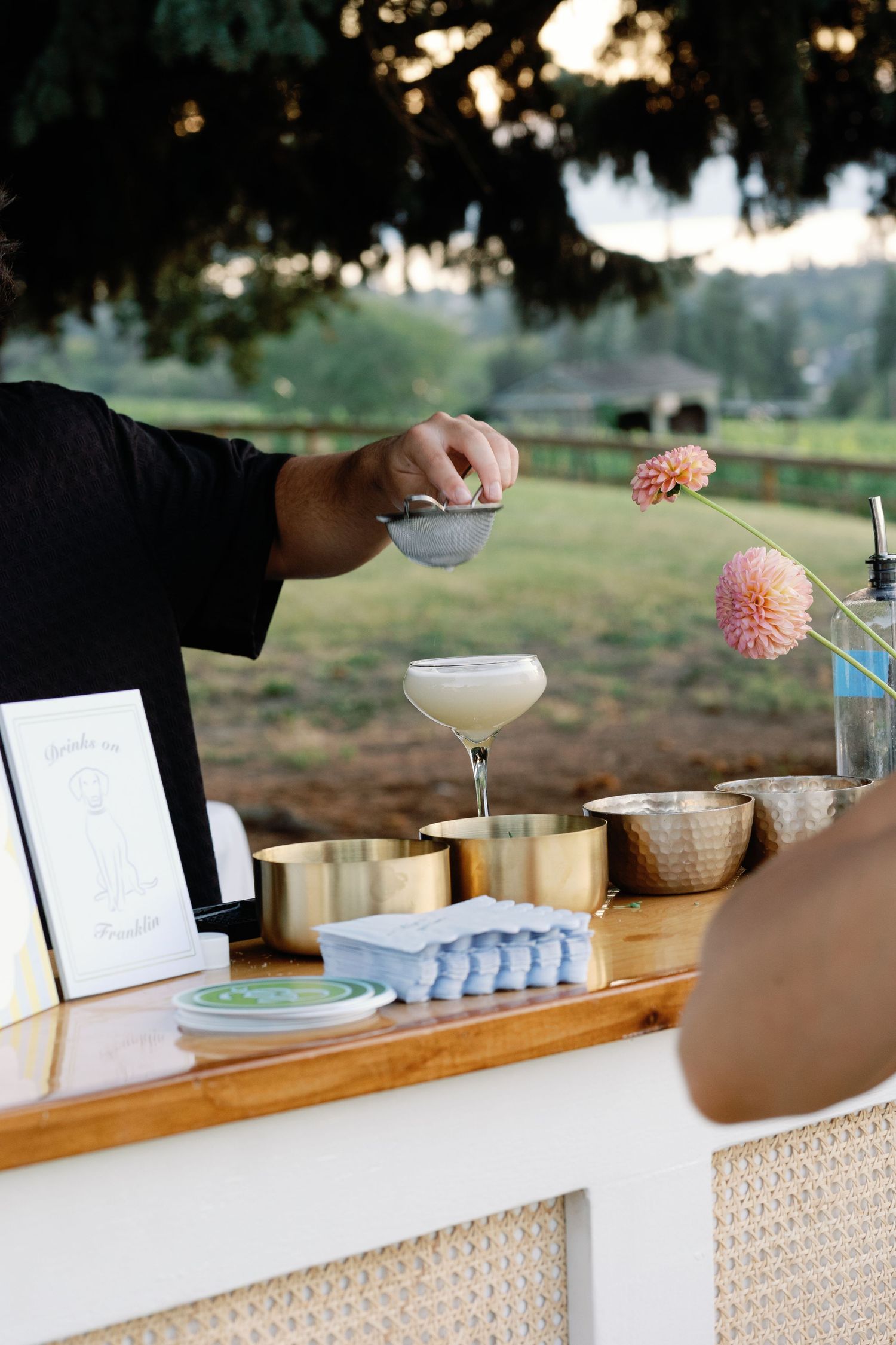 Bartender strains a cocktail into a coupe glass at an elegant outdoor wedding bar.