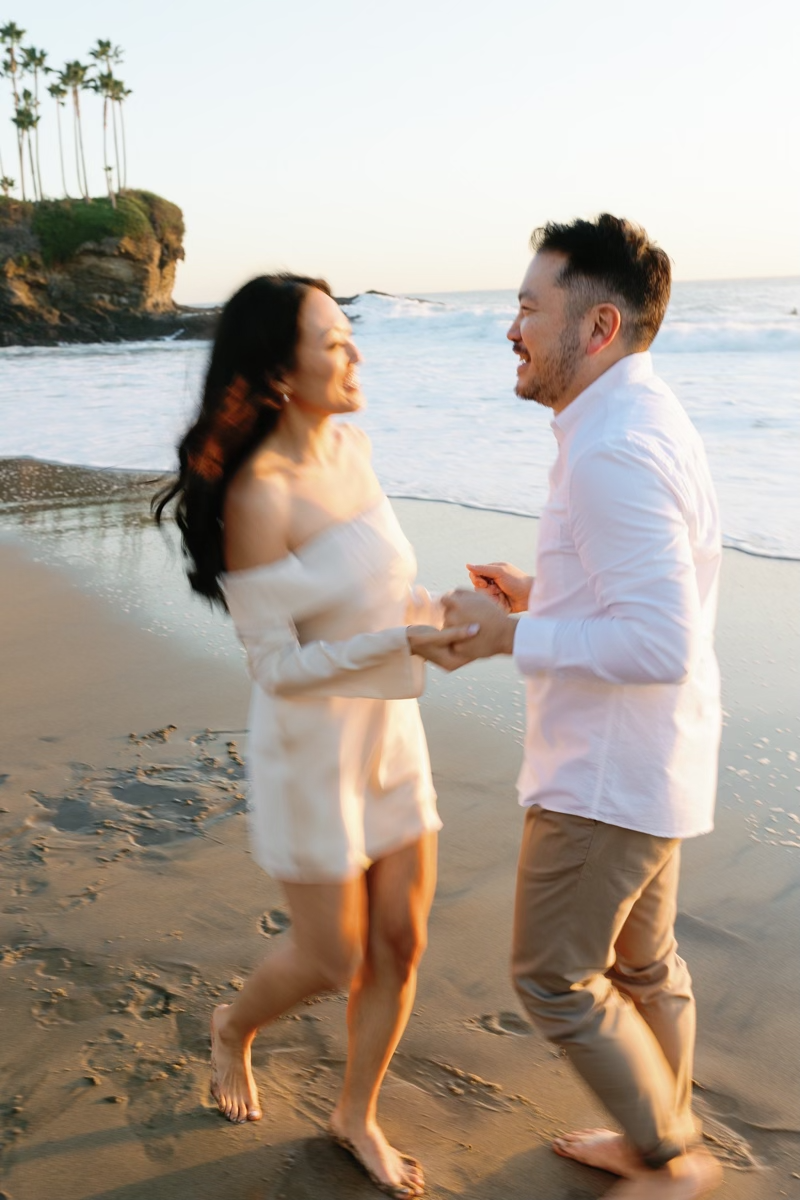 A laughing couple holds hands and dances together on a sandy beach at golden hour with ocean waves behind them.