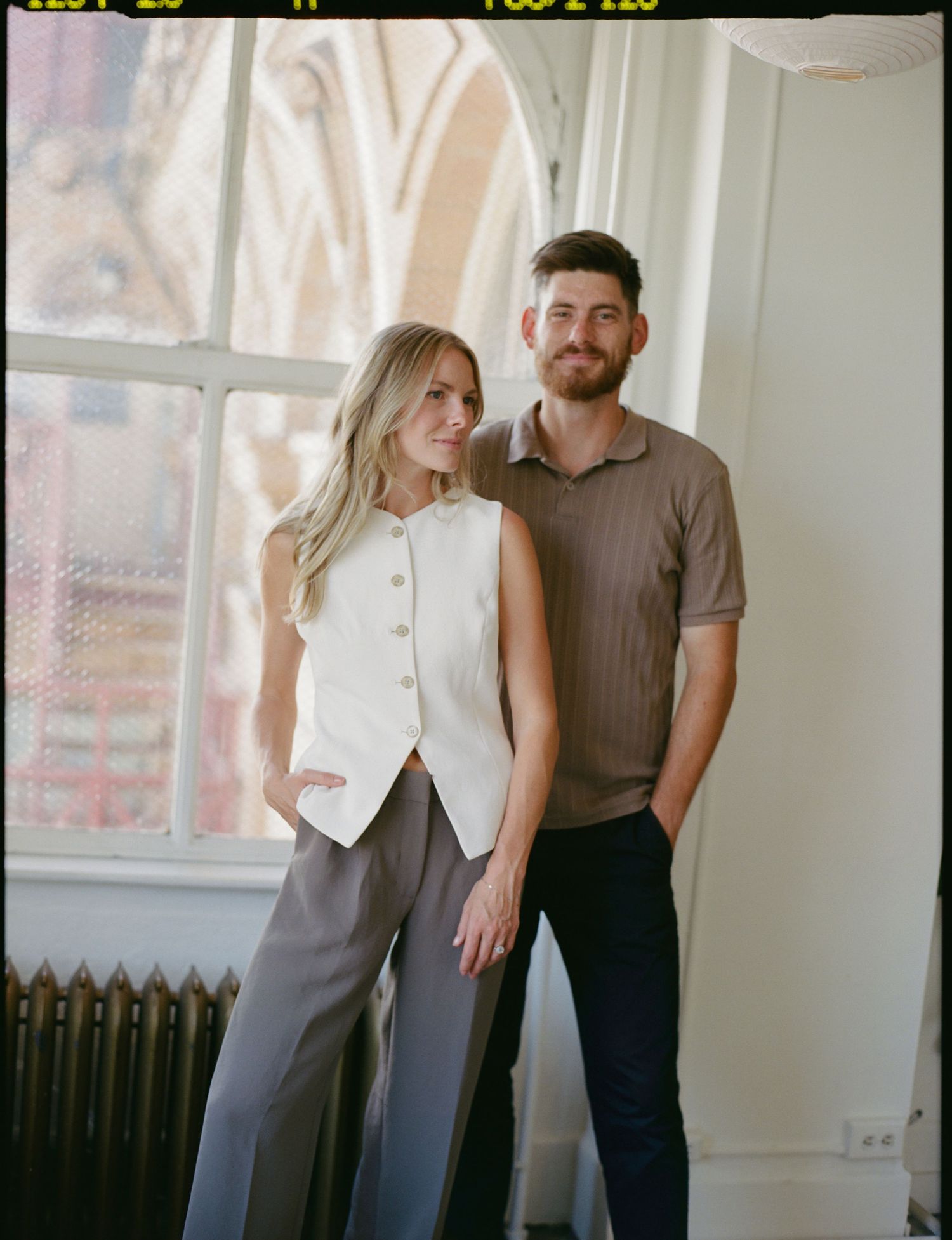 Stylish couple posing by a large window, woman in white vest and gray trousers, man in brown knit polo shirt.
