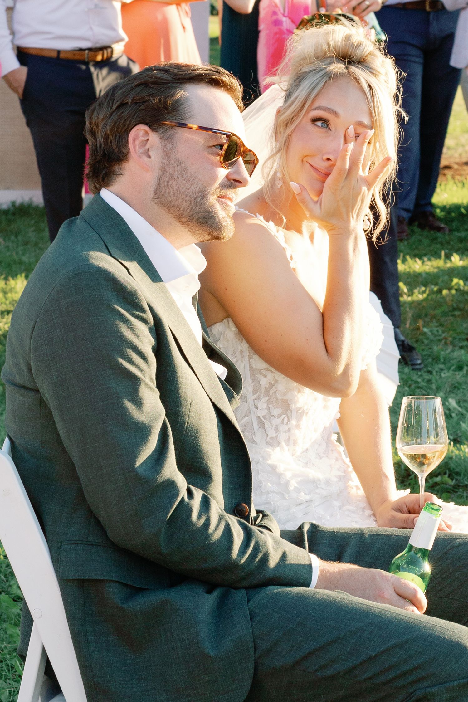 Emotional bride wipes tear while groom in green suit smiles during outdoor wedding reception.