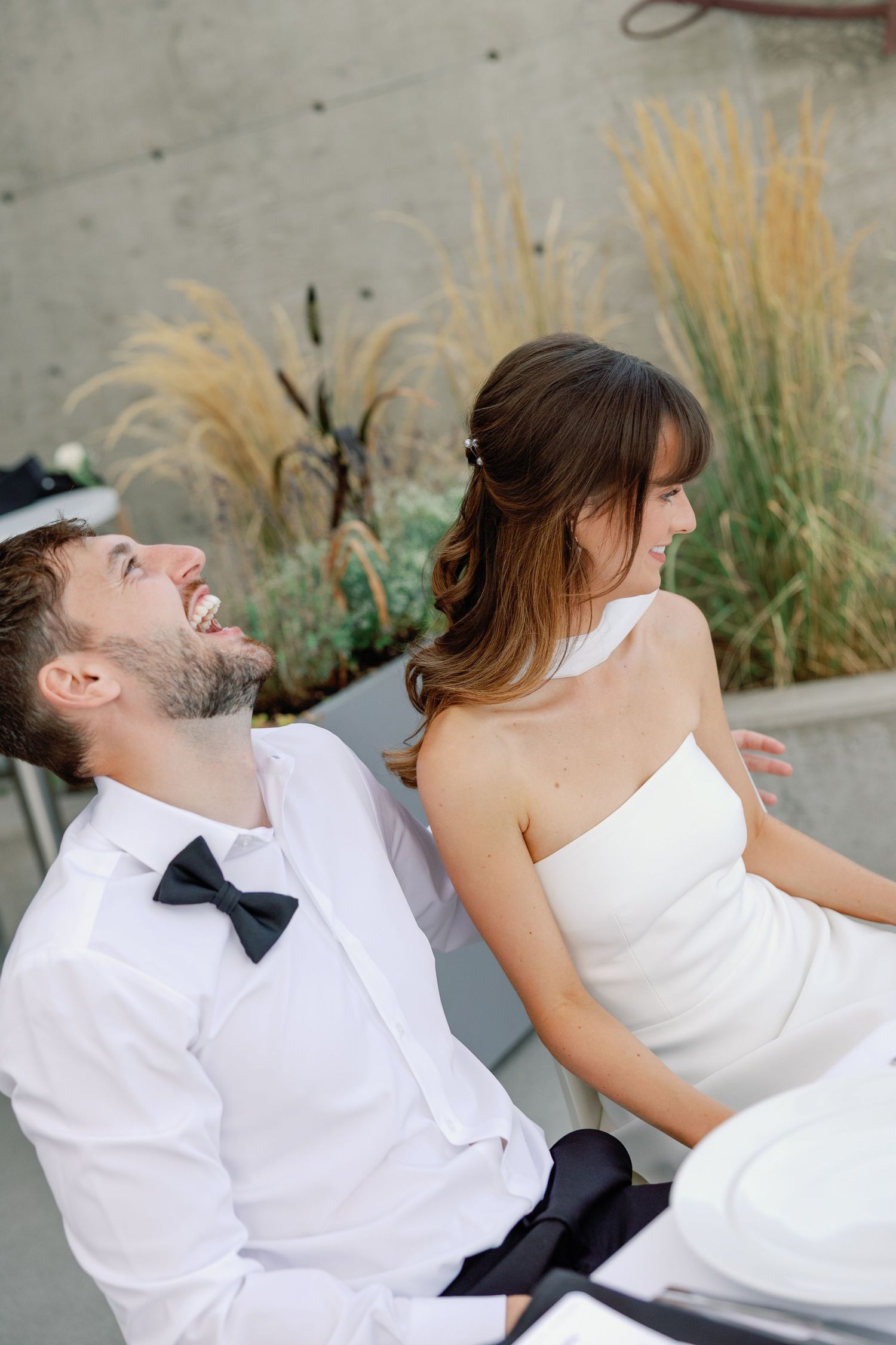Laughing bride and groom sharing a joyful moment seated outdoors with ornamental grasses in the background.