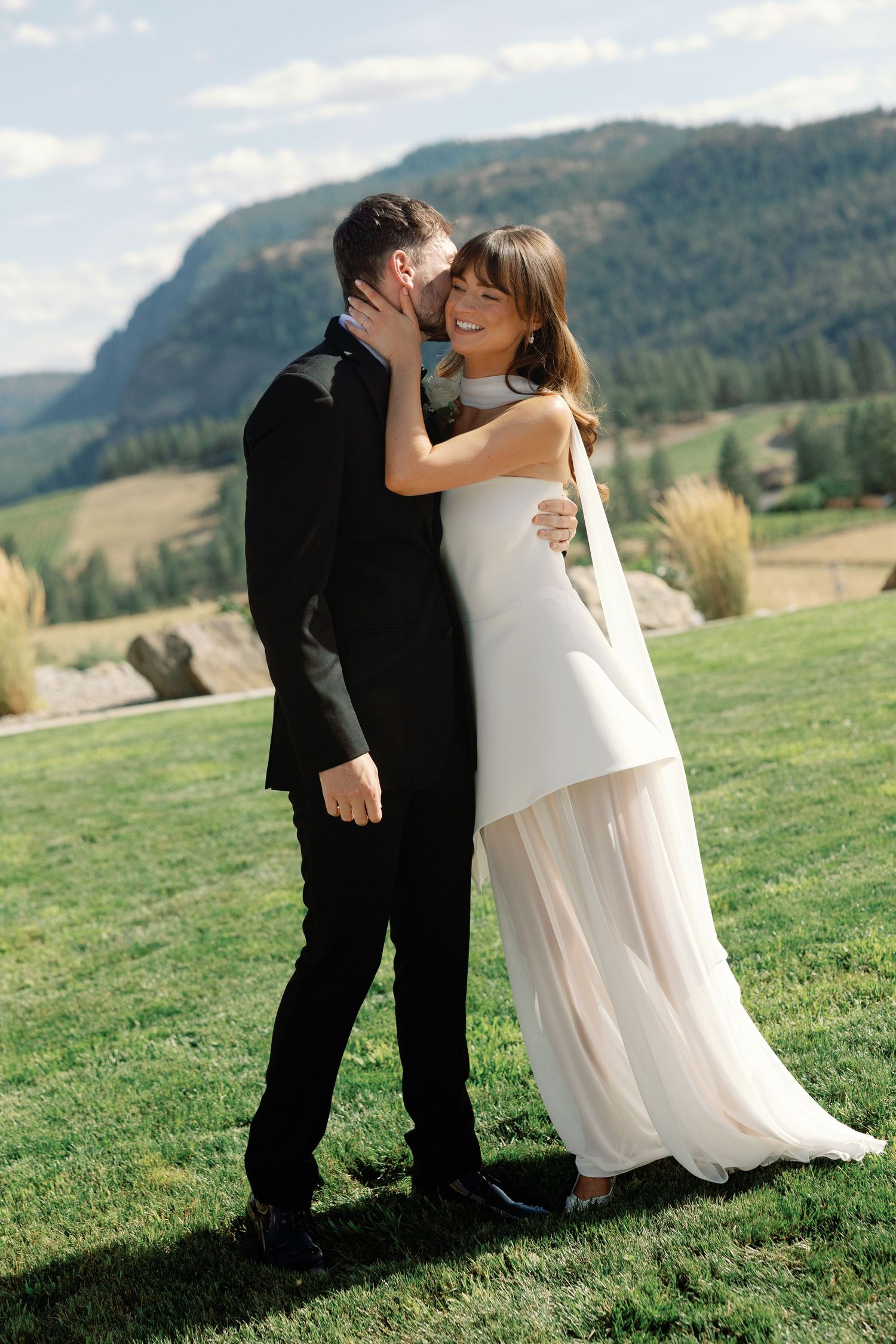Smiling bride in white layered gown embracing groom in black tuxedo with mountain scenery.