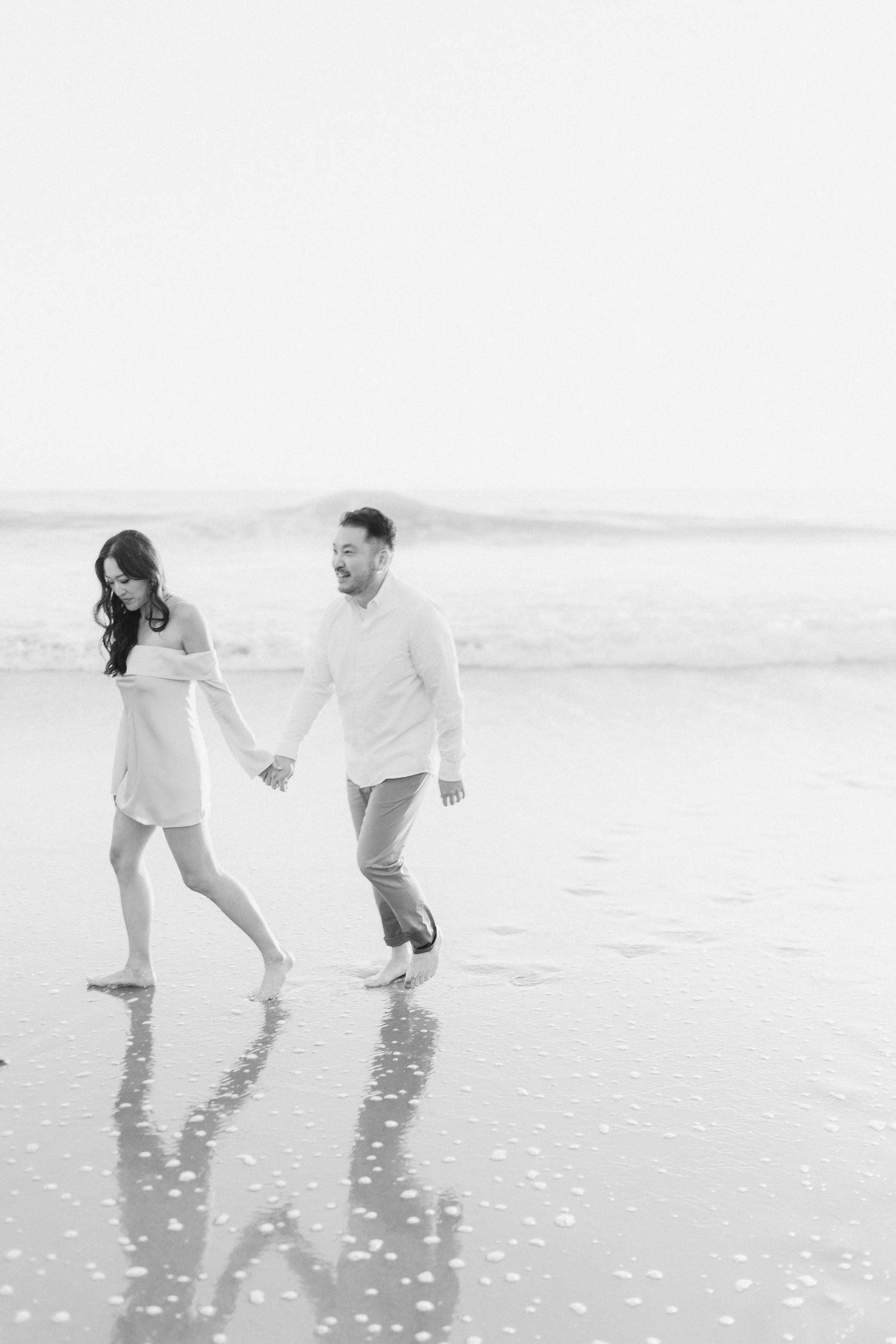 A black and white photo of a couple holding hands and walking barefoot along a reflective wet beach shoreline.
