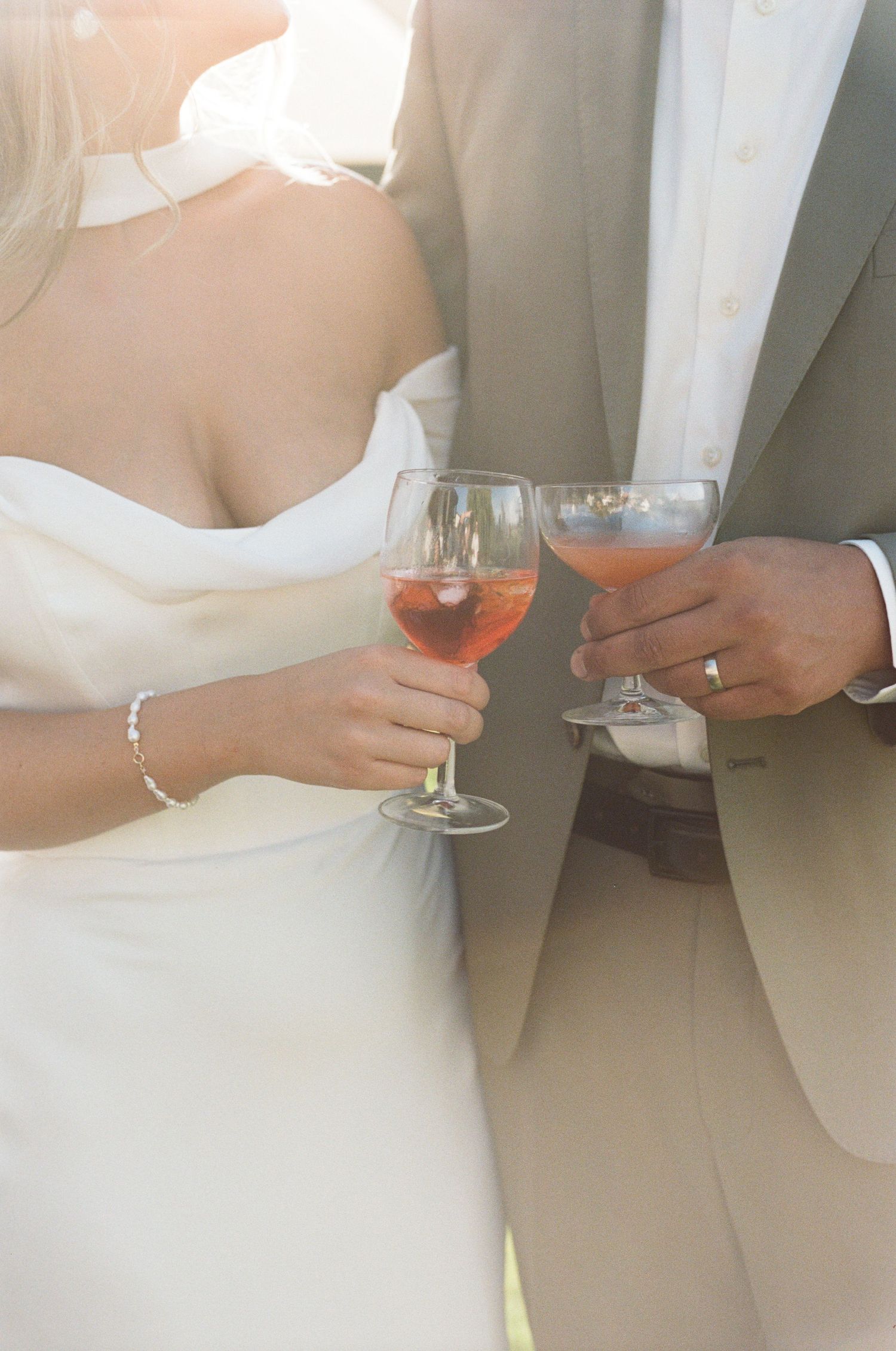 Bride and groom clink glasses of rose wine, both wearing wedding rings in warm golden sunlight.