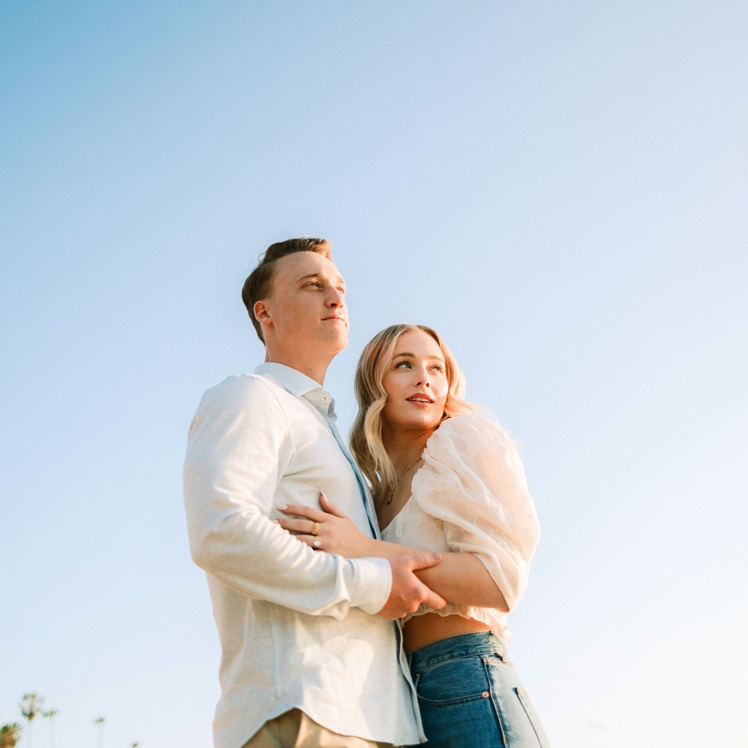 A couple embraces outdoors under a clear blue sky, the woman holding the man from behind in casual attire.