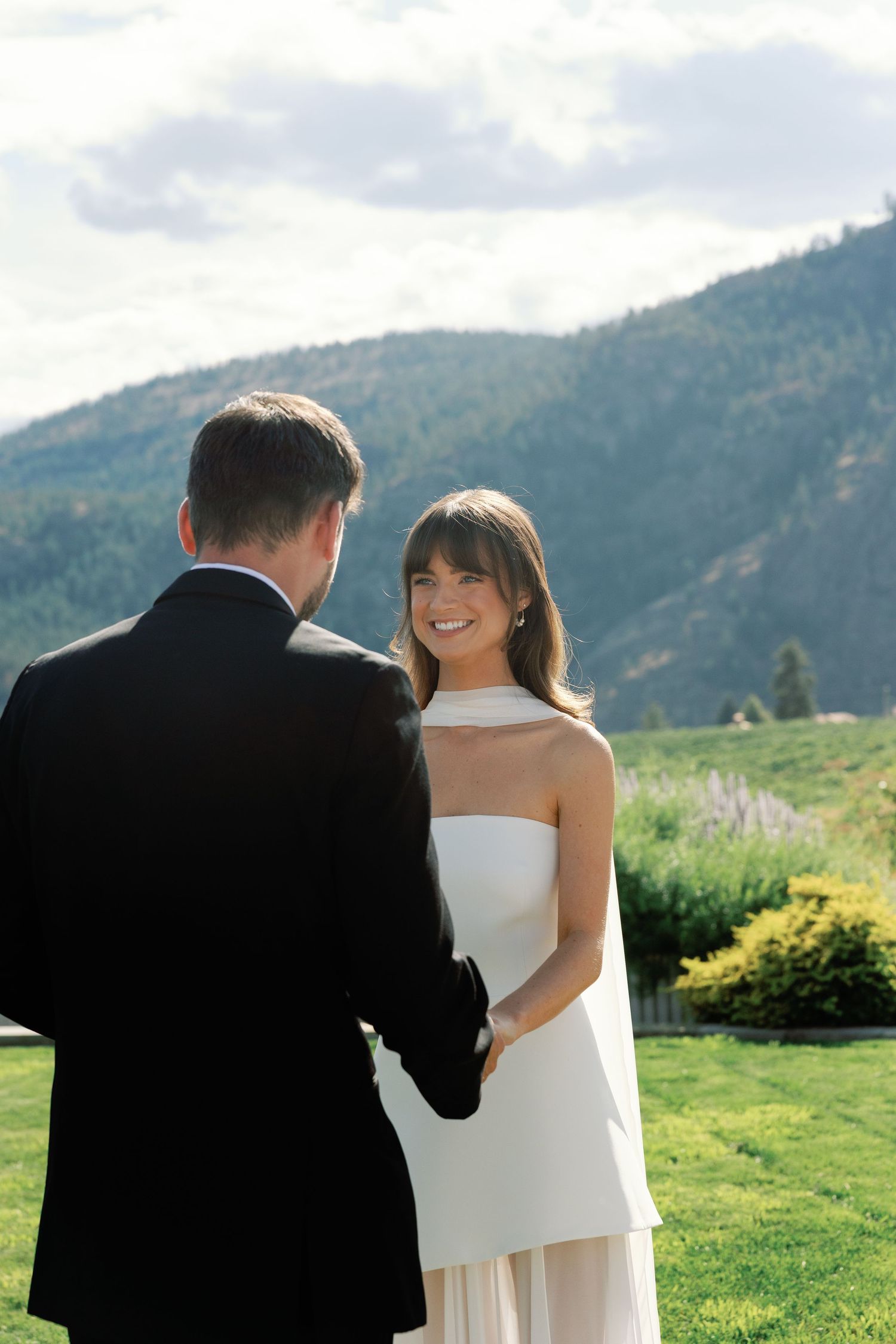 Bride in white halter gown smiles at groom during outdoor wedding ceremony with mountain views.