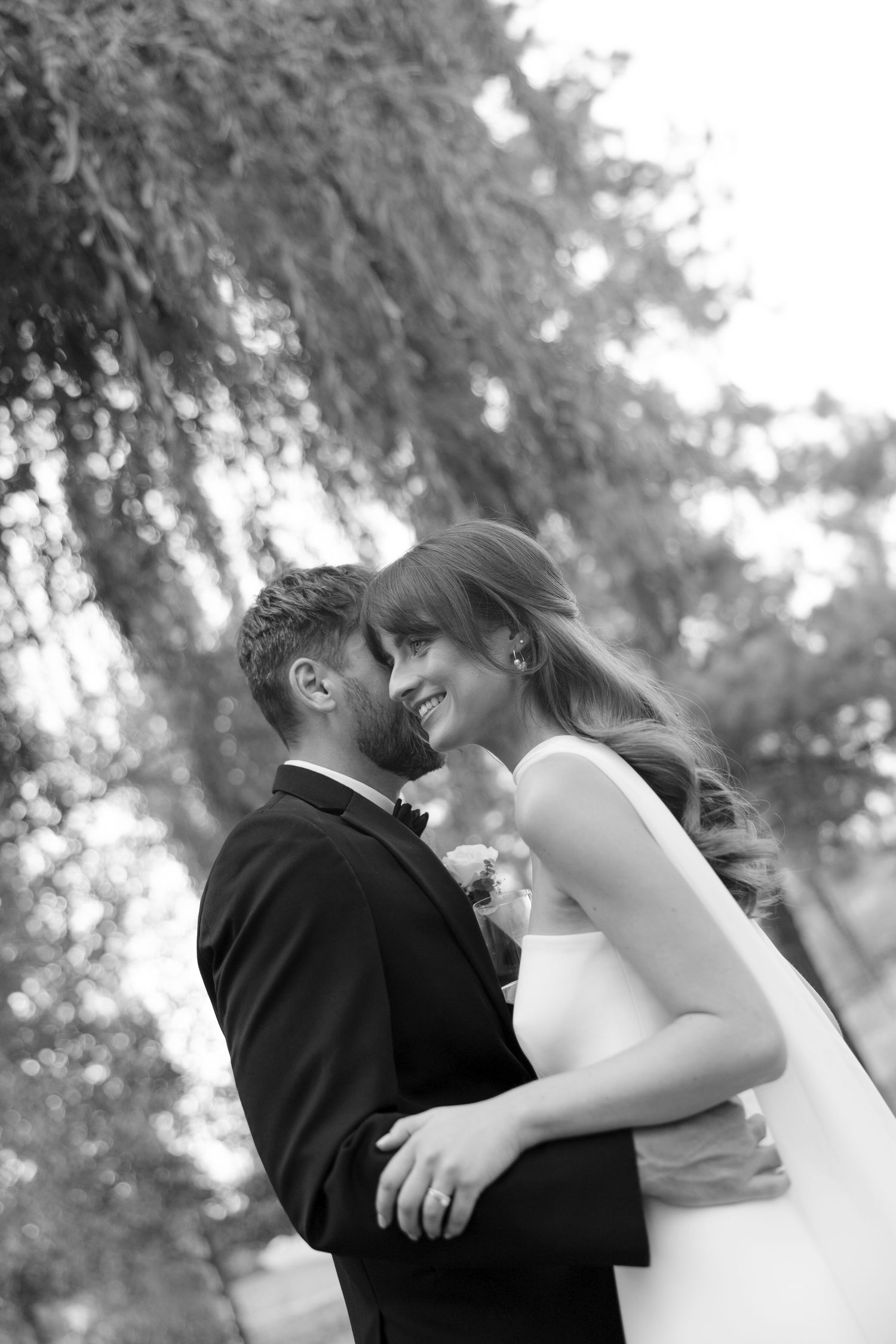 Black and white romantic portrait of smiling bride and groom embracing closely under a weeping willow tree.