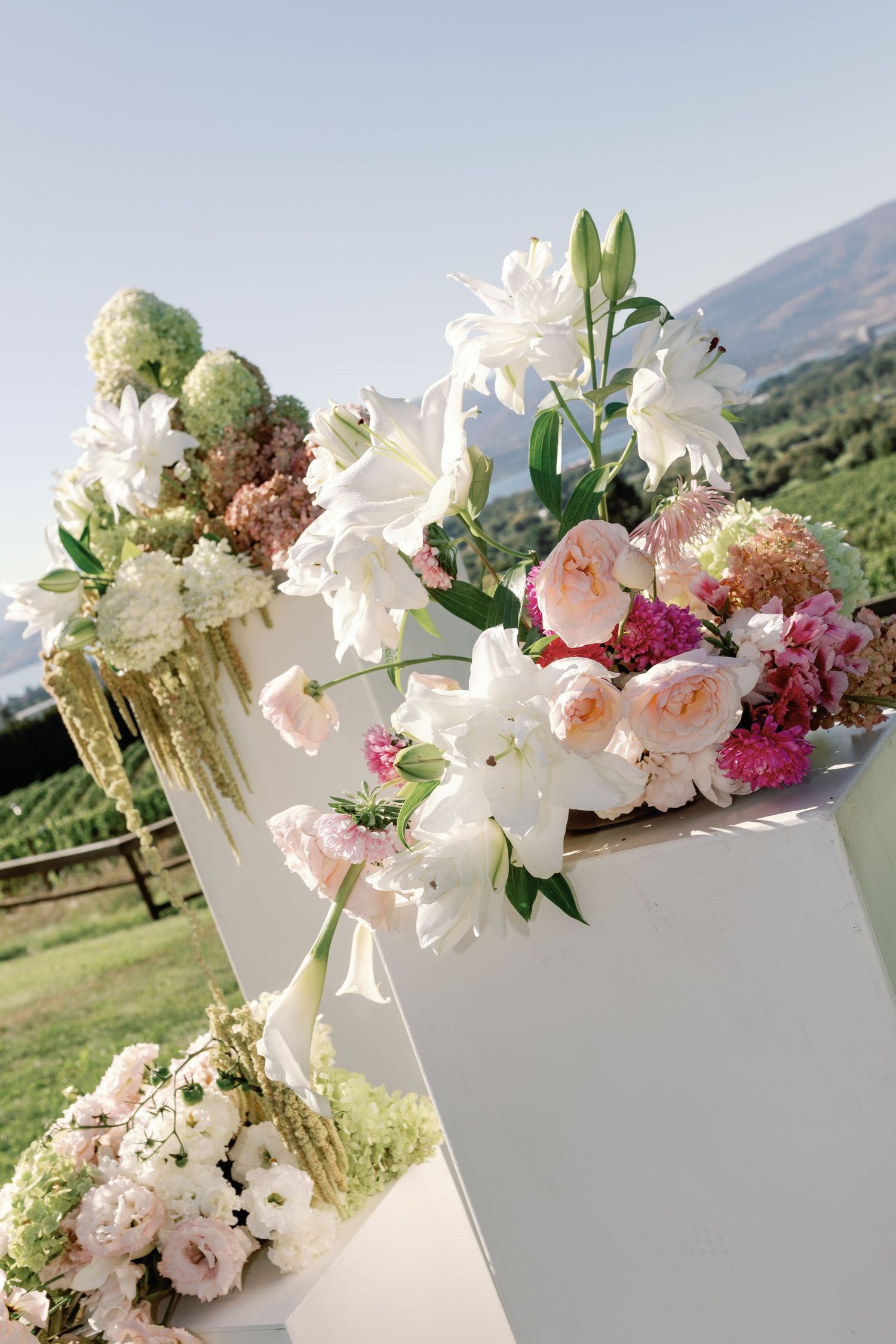 Lush wedding ceremony arch adorned with white lilies, pink roses, and hydrangeas against mountain vineyard backdrop.