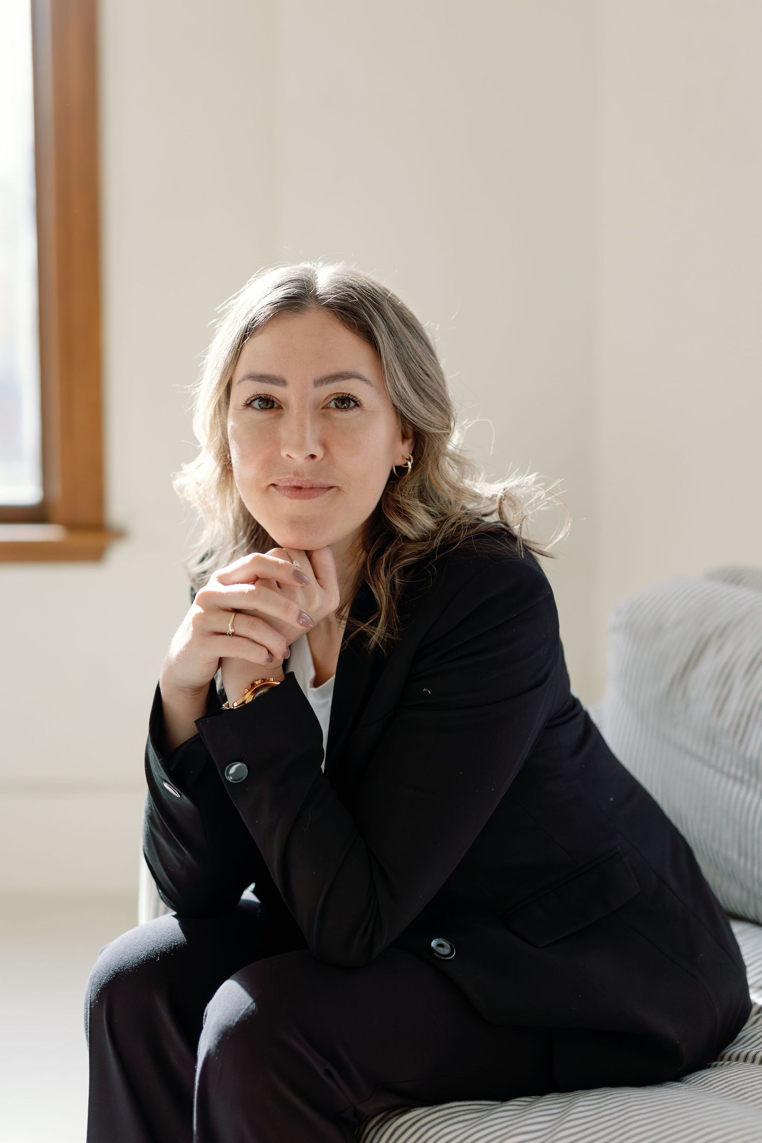A professional woman in a black blazer sits thoughtfully with hands clasped, posing in a bright modern interior.