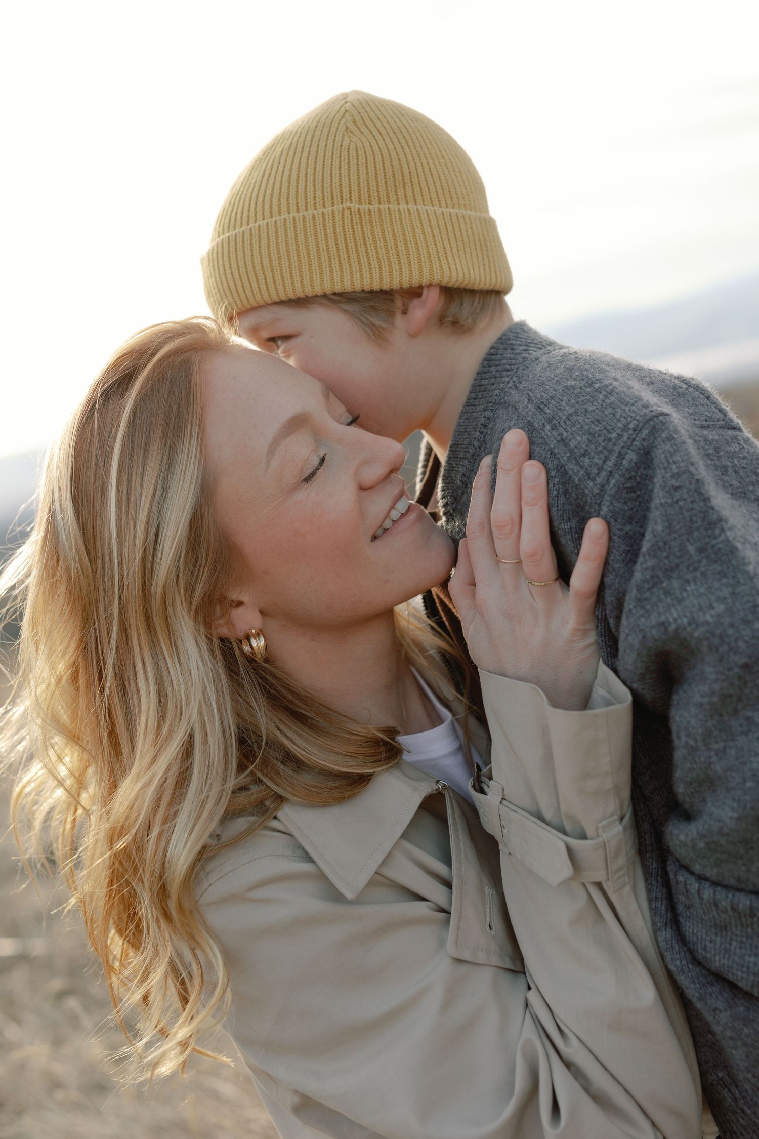 A mother and young child share a tender kiss outdoors, with golden sunlight illuminating their warm embrace.