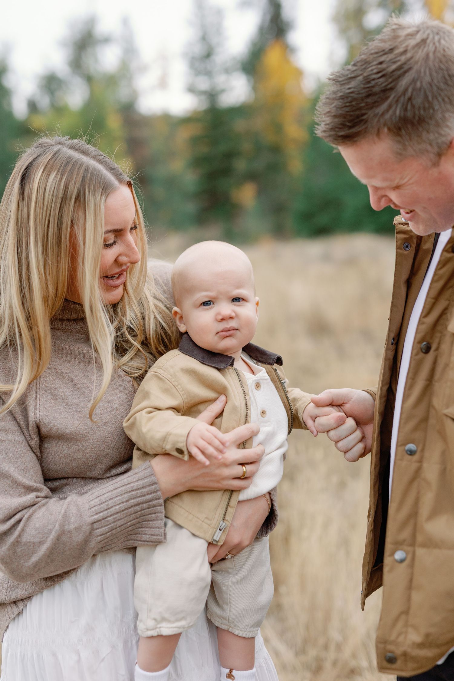 A couple smiles at their baby outdoors in autumn, dressed in neutral tones with colorful fall trees in the background.