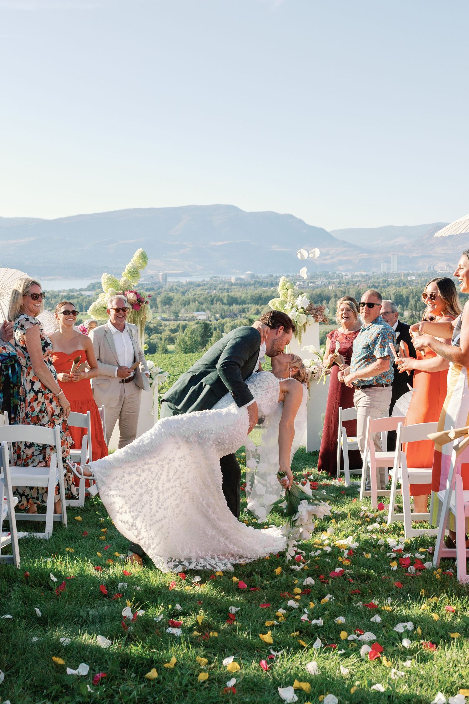 Bride and groom share a romantic dip kiss at outdoor wedding ceremony with mountain lake views.
