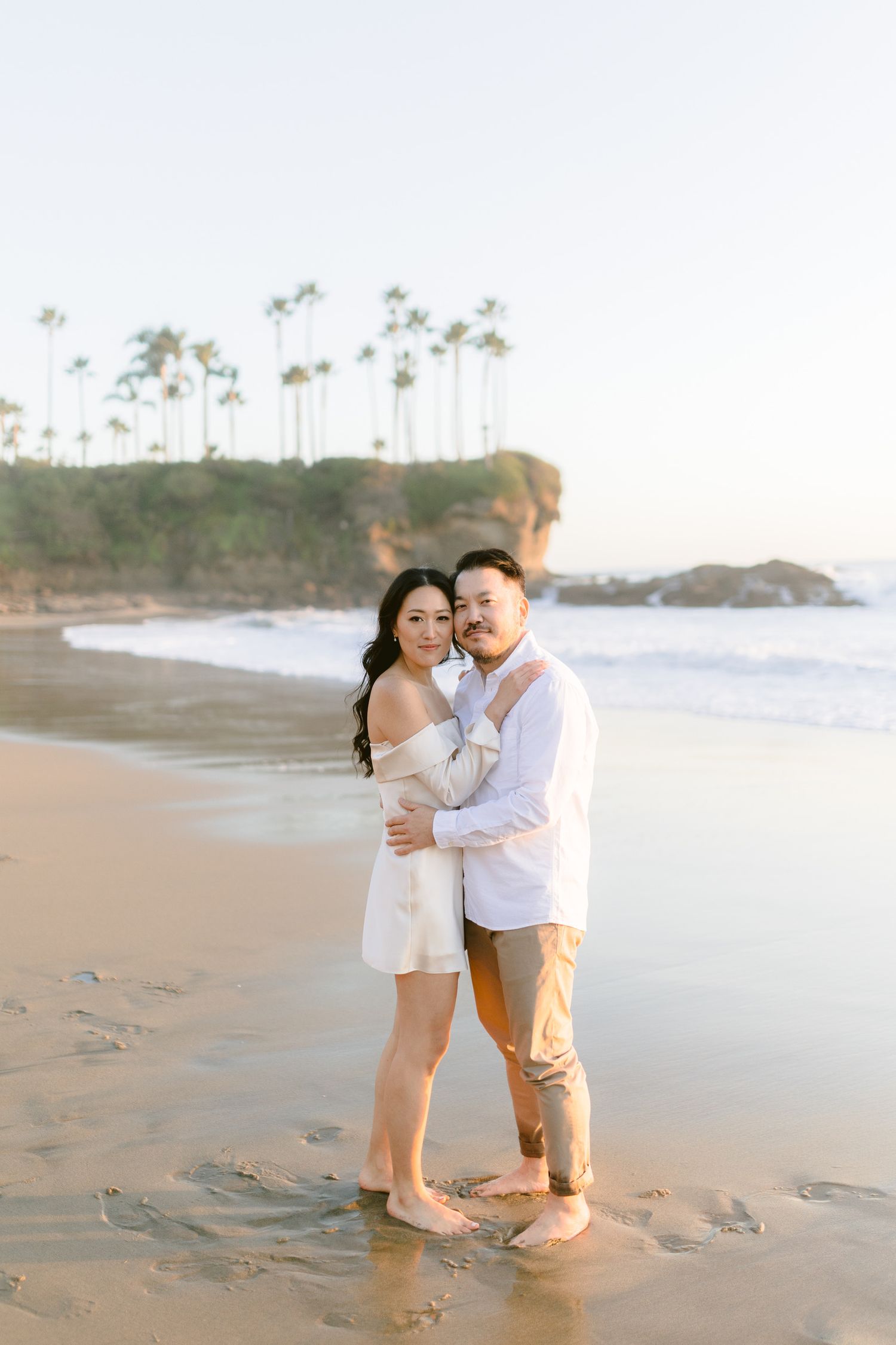 A couple embraces barefoot on a sandy beach at golden hour with palm trees and cliffs in the background.