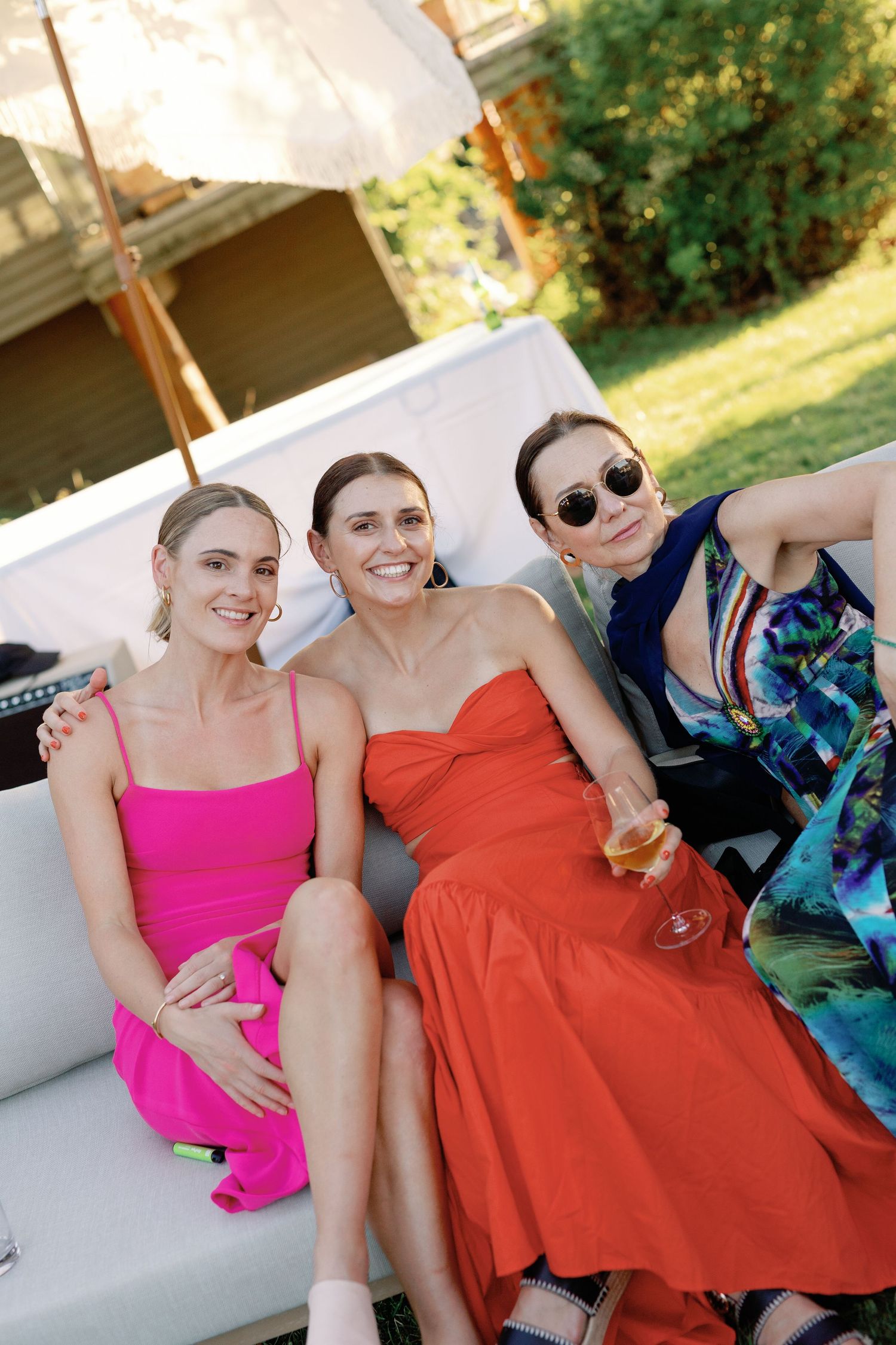 Three women in pink, orange, and blue dresses smile together on outdoor wedding lounge seating.