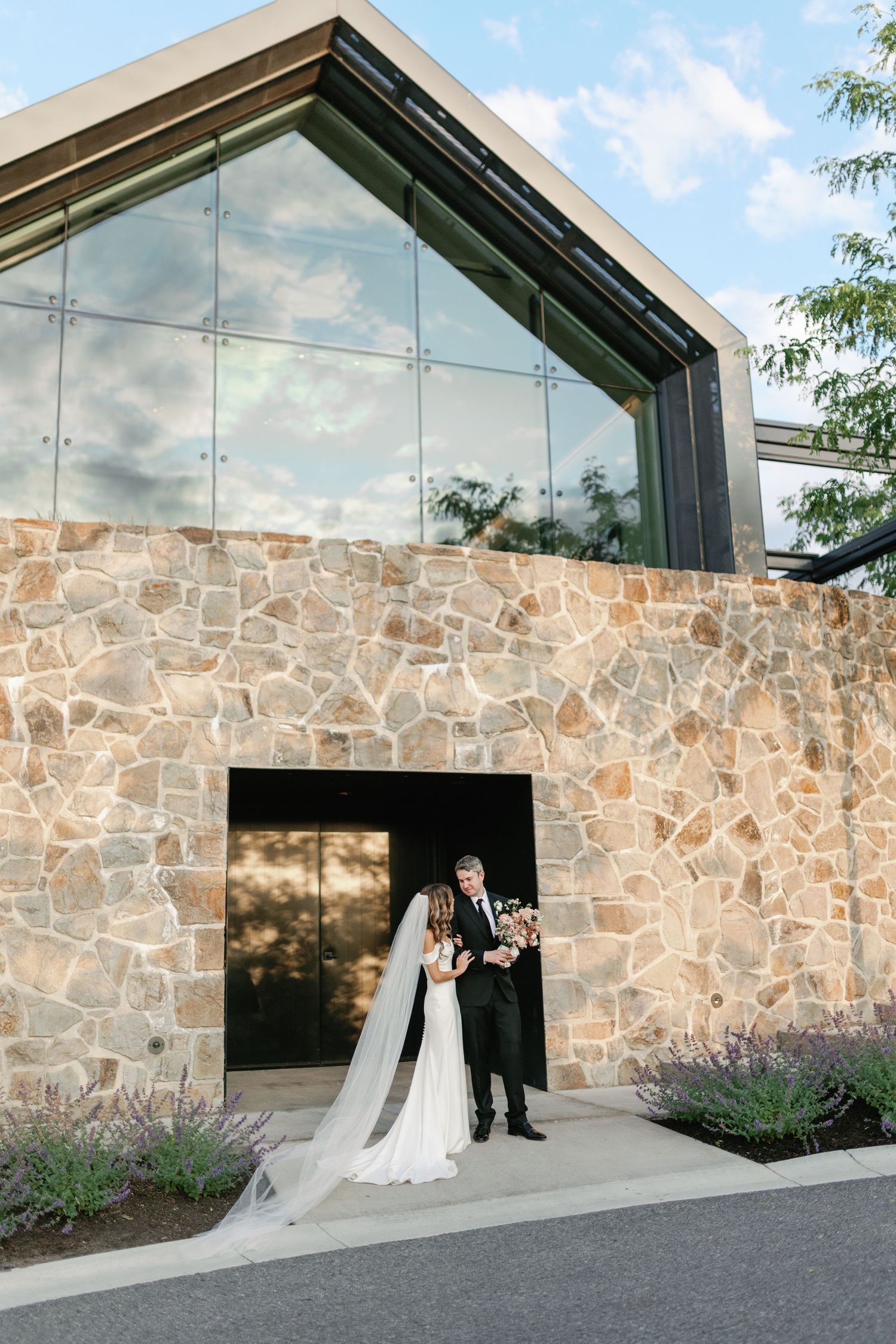 Bride and groom posing before modern stone and glass wedding venue at golden hour.