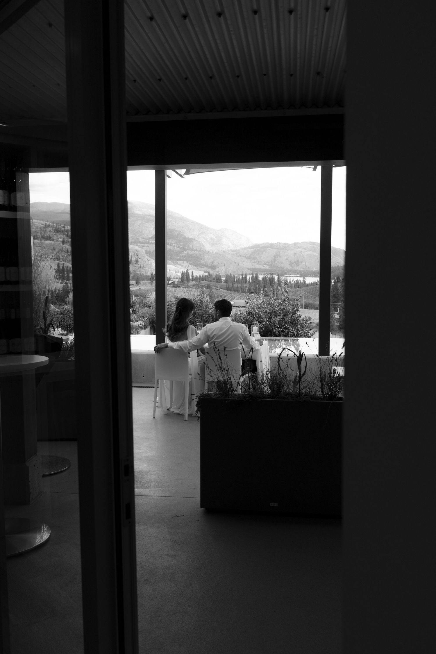 Black and white photo of a couple standing together gazing at a scenic mountain valley view through large windows.