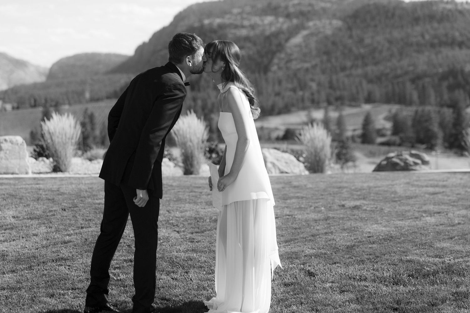 Black and white photo of bride and groom sharing first kiss at outdoor wedding.