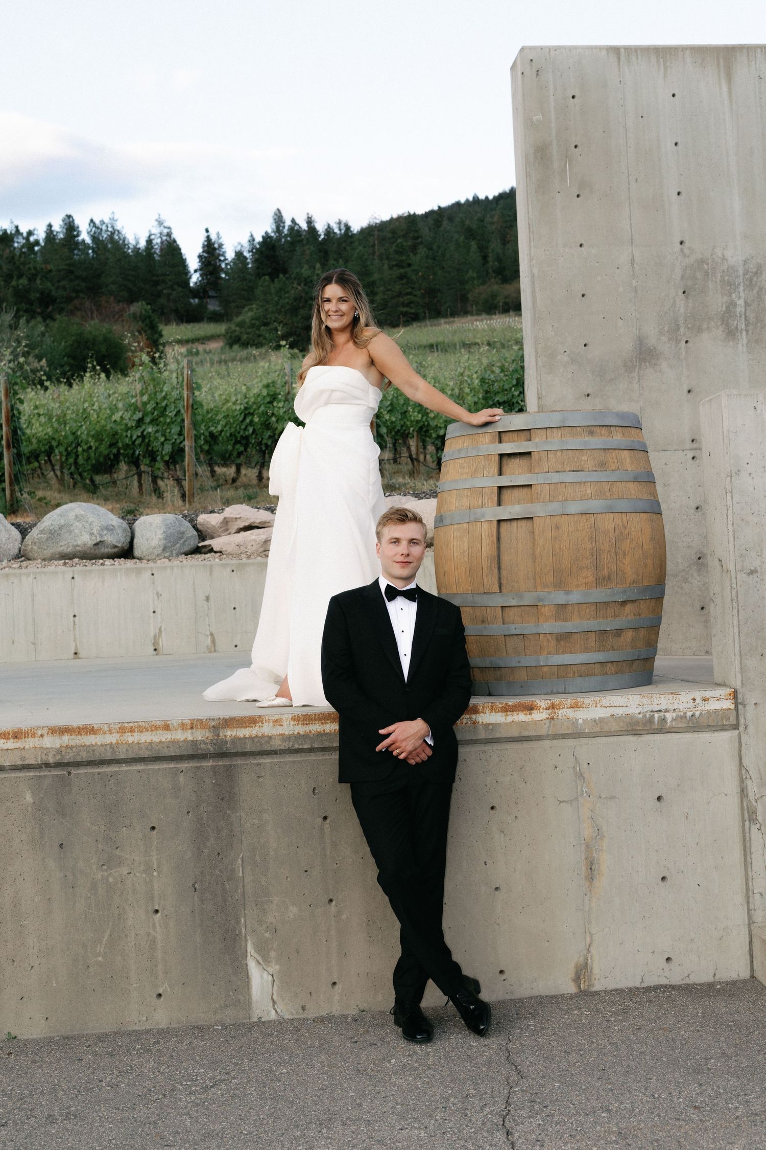 Bride in white gown stands on ledge near wine barrel as groom in tuxedo leans against wall at vineyard wedding.