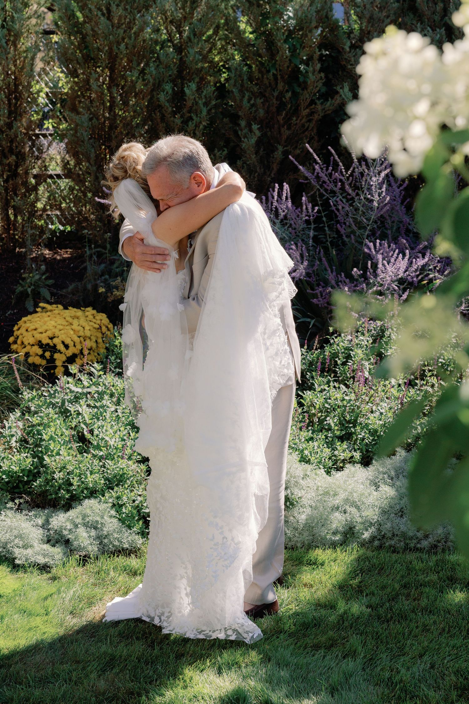 Bride in lace gown and veil shares emotional hug with older man in lush garden during first look.