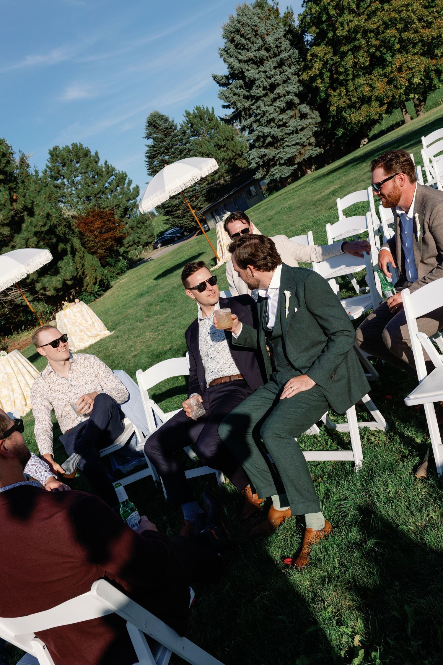 Groomsmen in suits laugh and chat on white chairs at sunny outdoor wedding ceremony.
