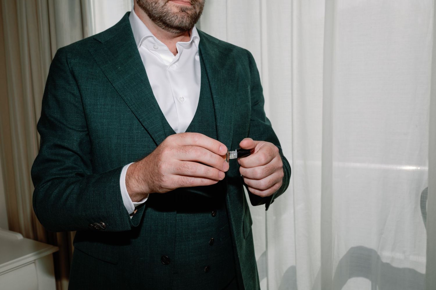 Groom in dark green plaid suit jacket fastens button while preparing for wedding ceremony indoors.