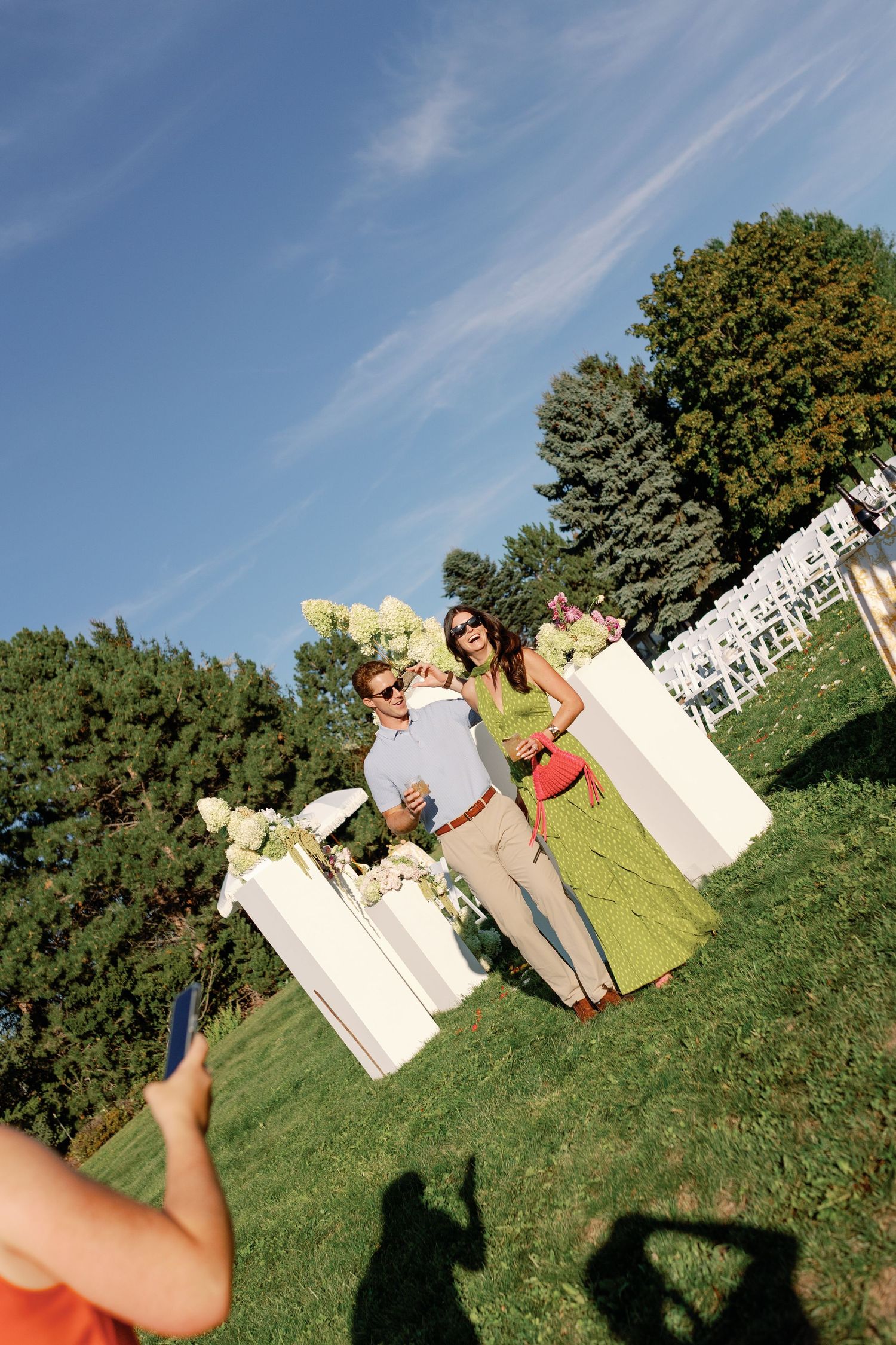 Wedding guests posing for photos beside white floral ceremony pillars on a sunny outdoor lawn.