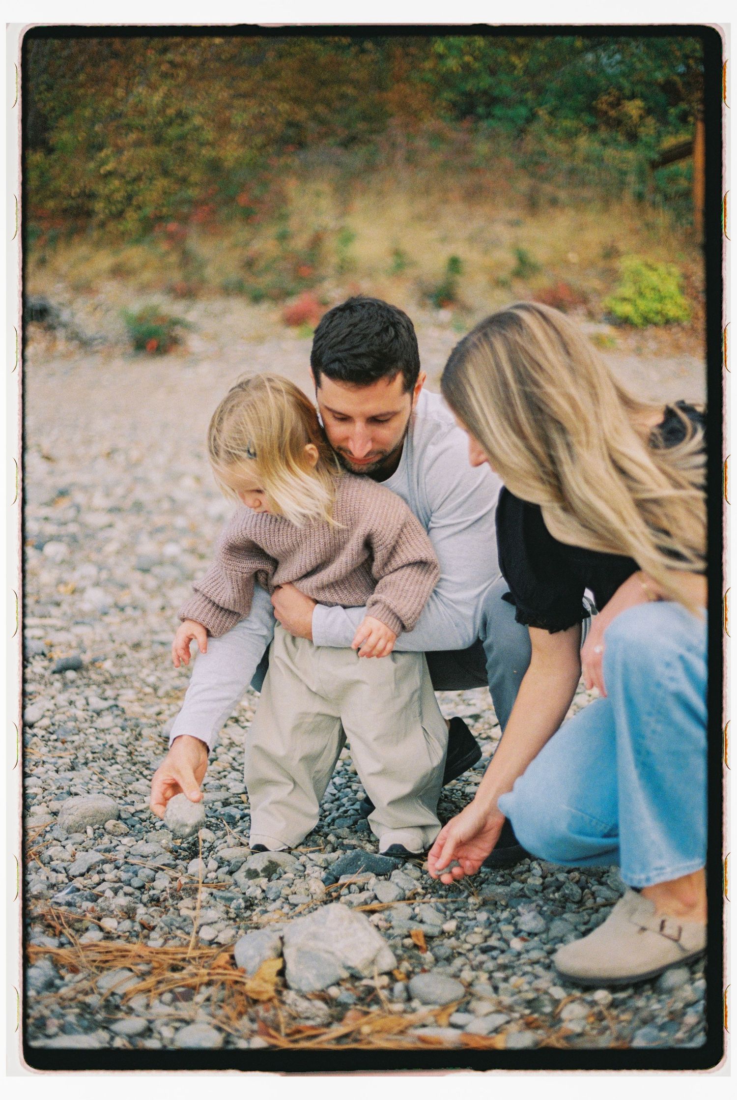 A family of three examines pebbles on a rocky beach, with parents crouching beside their young blonde child.