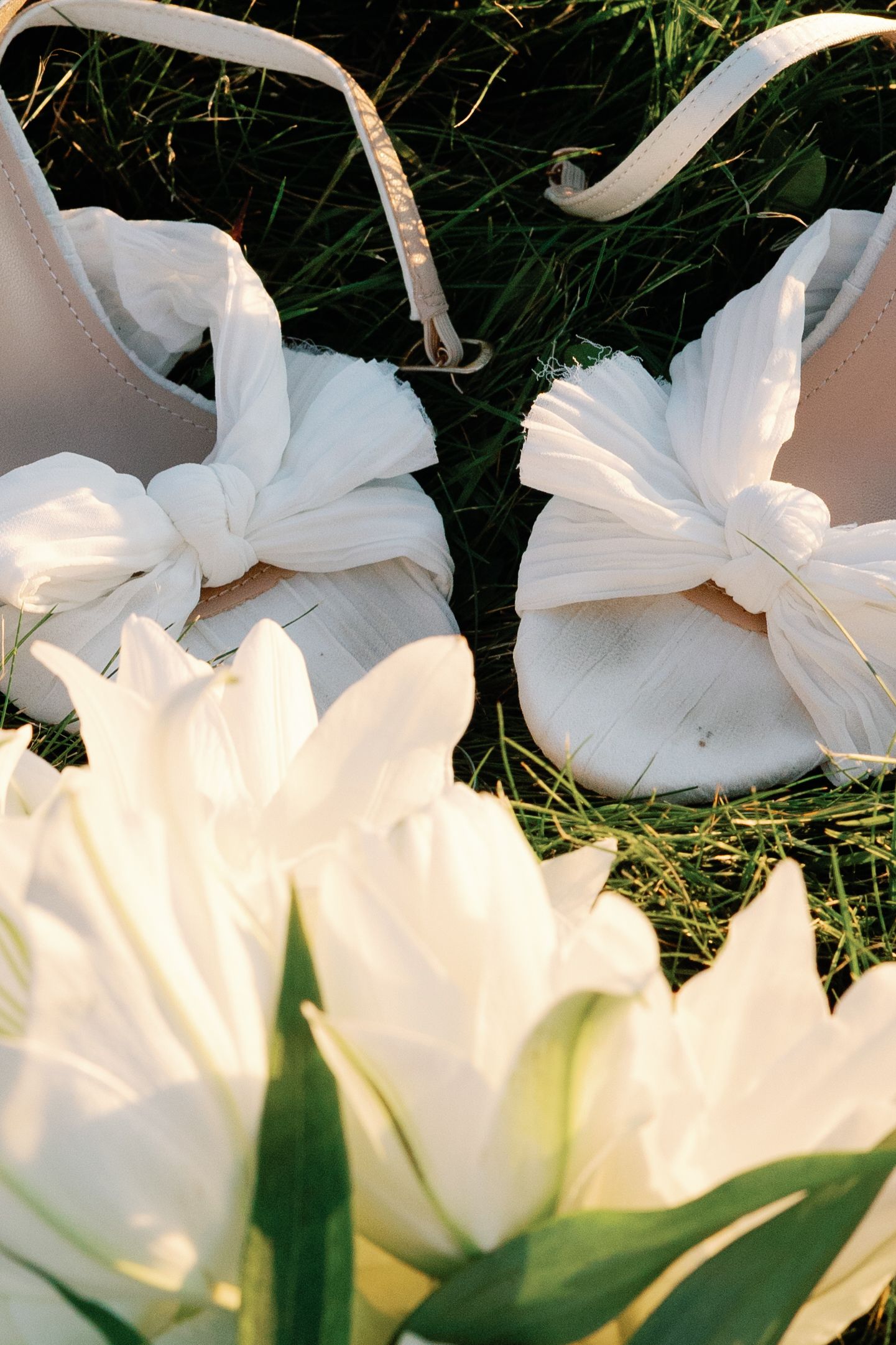 Close-up of white bow-detail bridal sandals on green grass surrounded by white tulips.