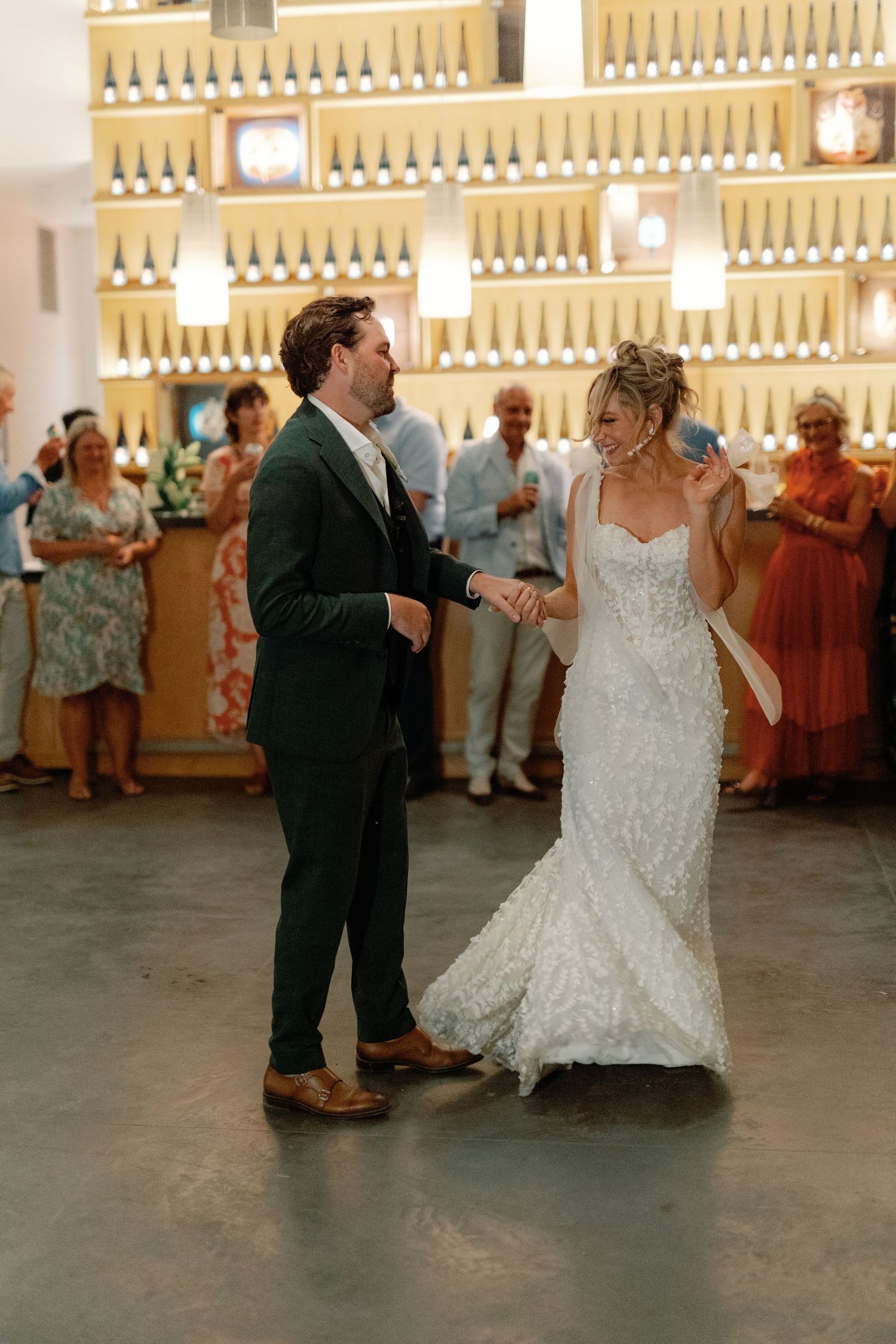 Bride and groom sharing first dance in winery venue with illuminated wine bottle display wall backdrop.