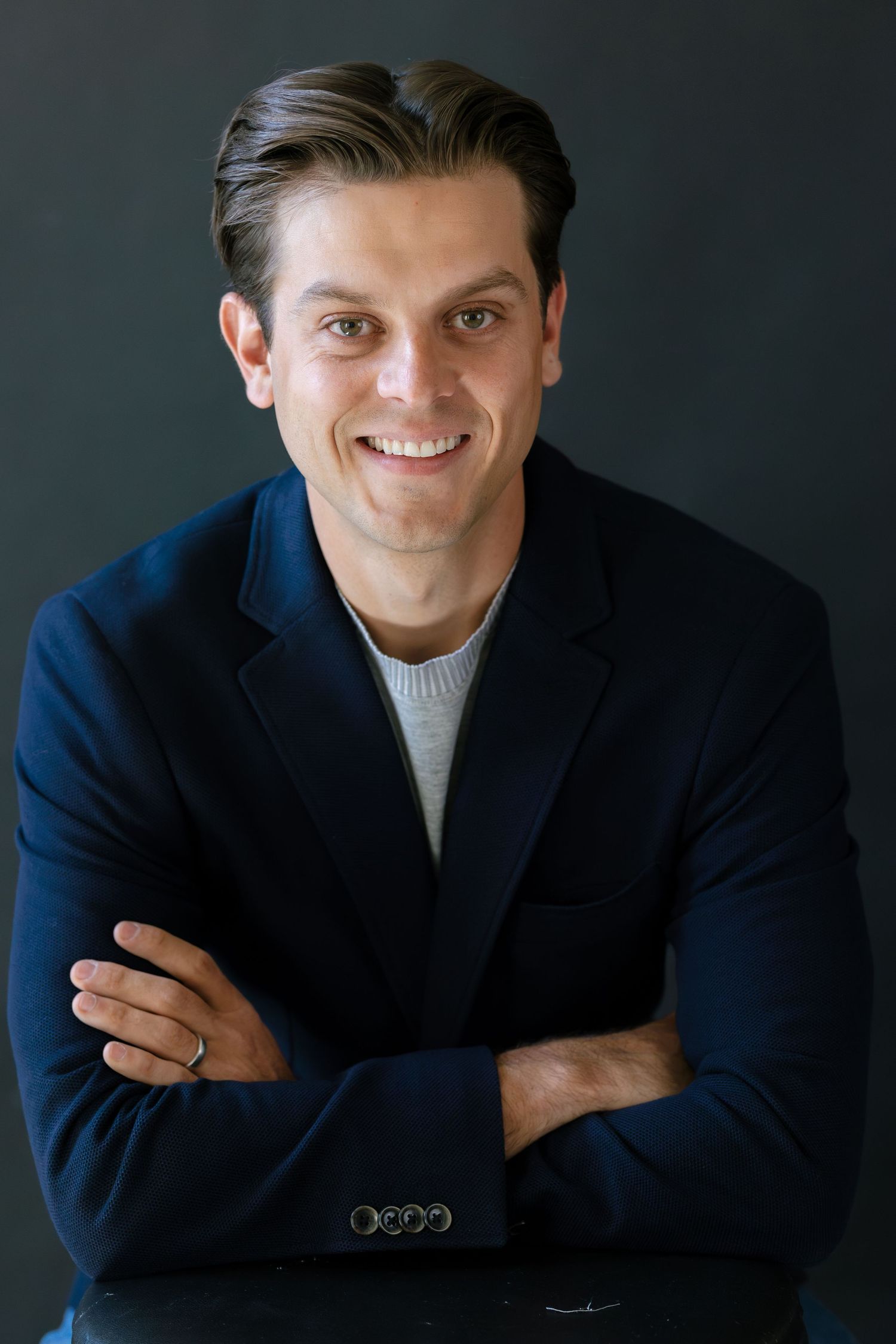 Smiling man in navy blazer and gray sweater with arms crossed poses confidently against dark background.