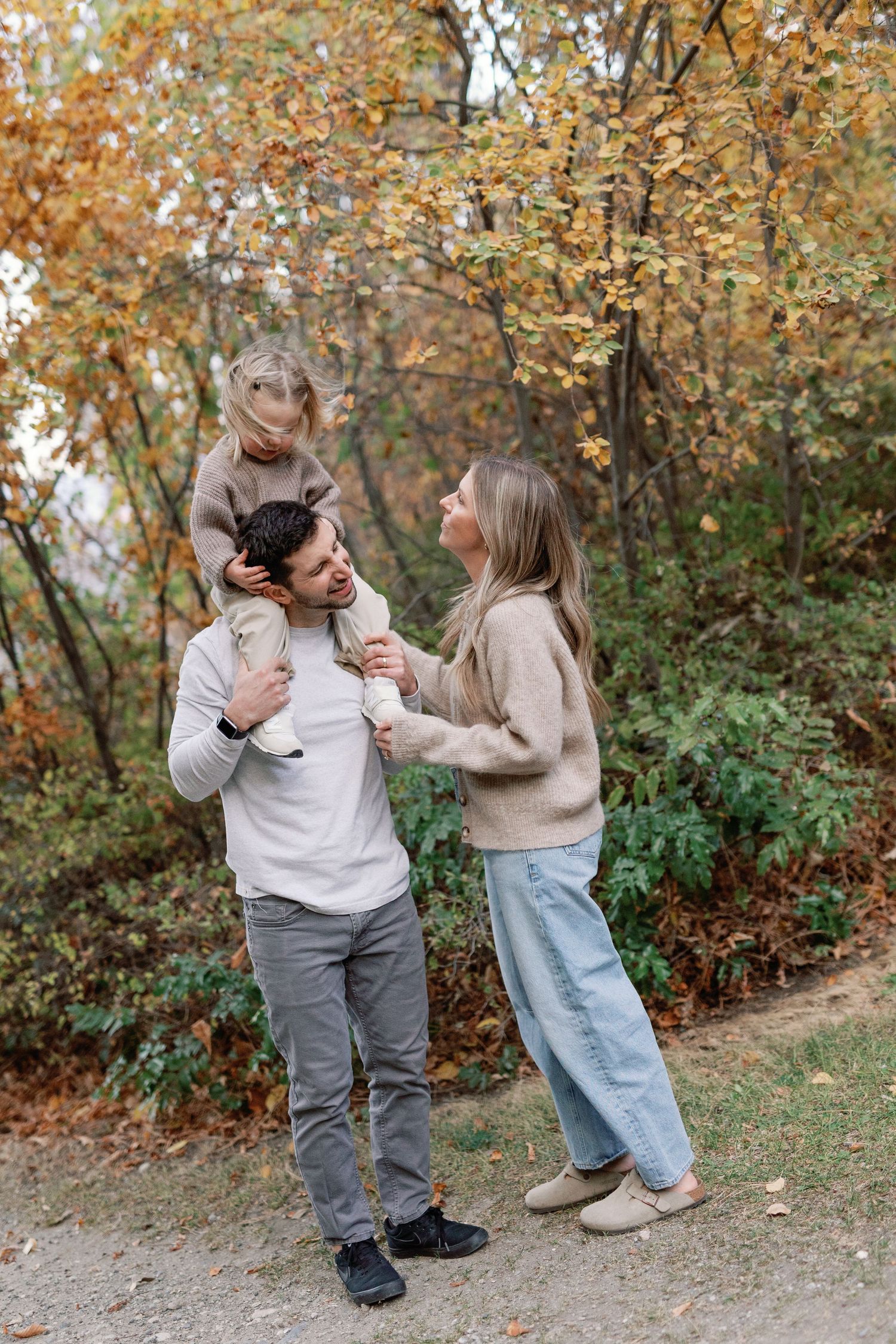 A happy family of three enjoys an autumn walk, with a child sitting on dad's shoulders while mom smiles in a fall park.