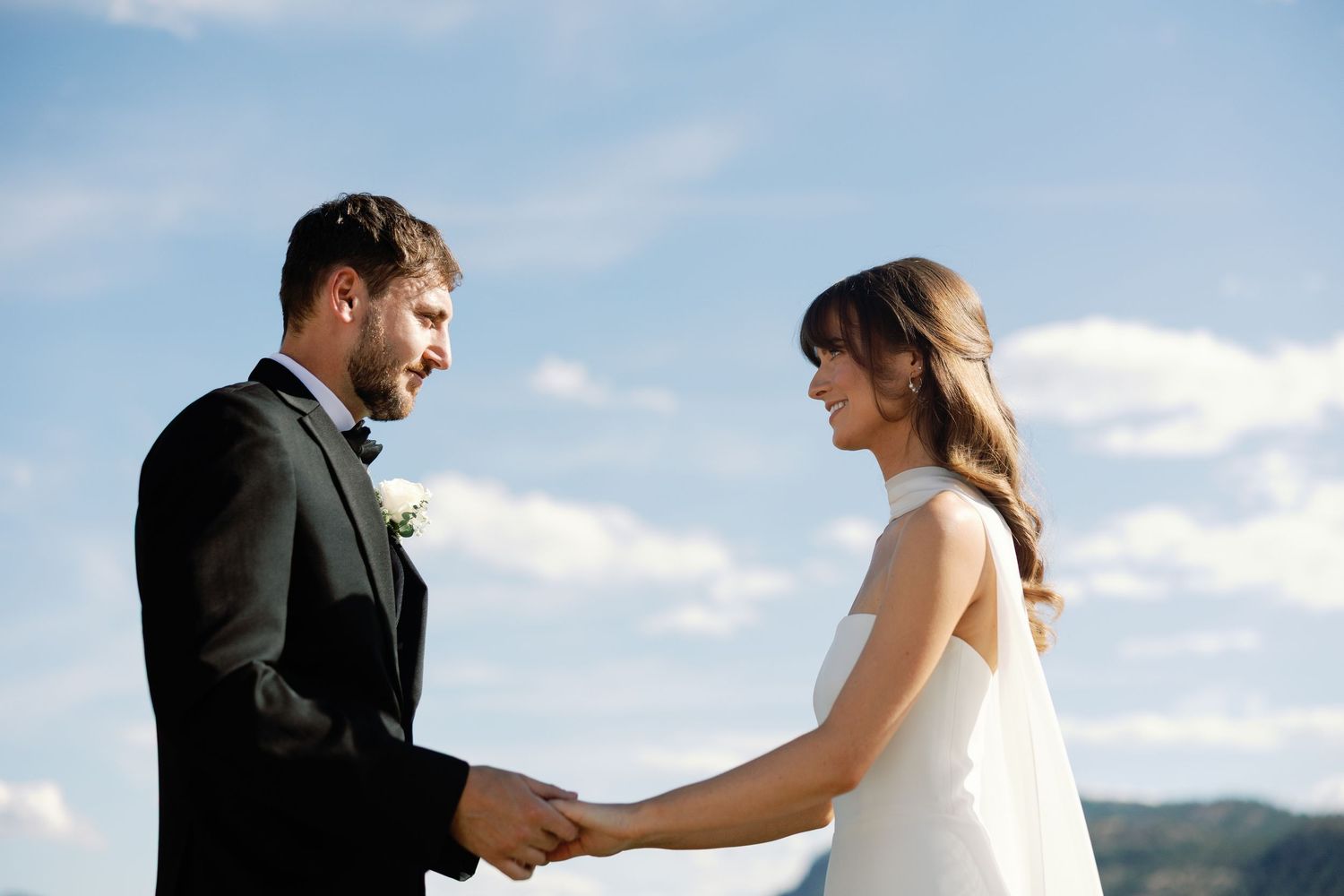 Smiling bride and groom hold hands under a bright blue sky during their outdoor wedding ceremony.