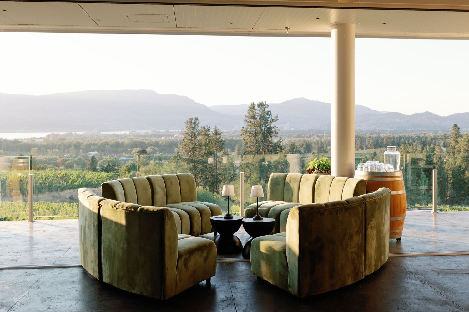 Elegant winery lounge with green velvet curved sofas overlooking vineyard and mountain views through floor-to-ceiling windows.