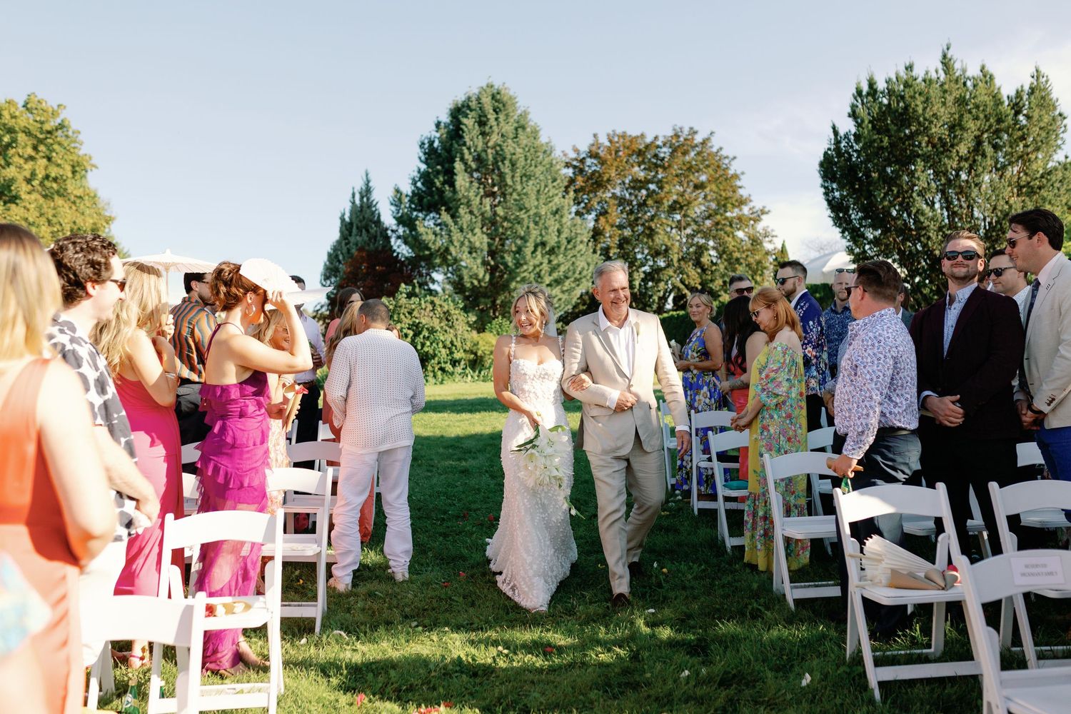 Bride and groom walking down aisle at outdoor garden wedding ceremony with white chairs.
