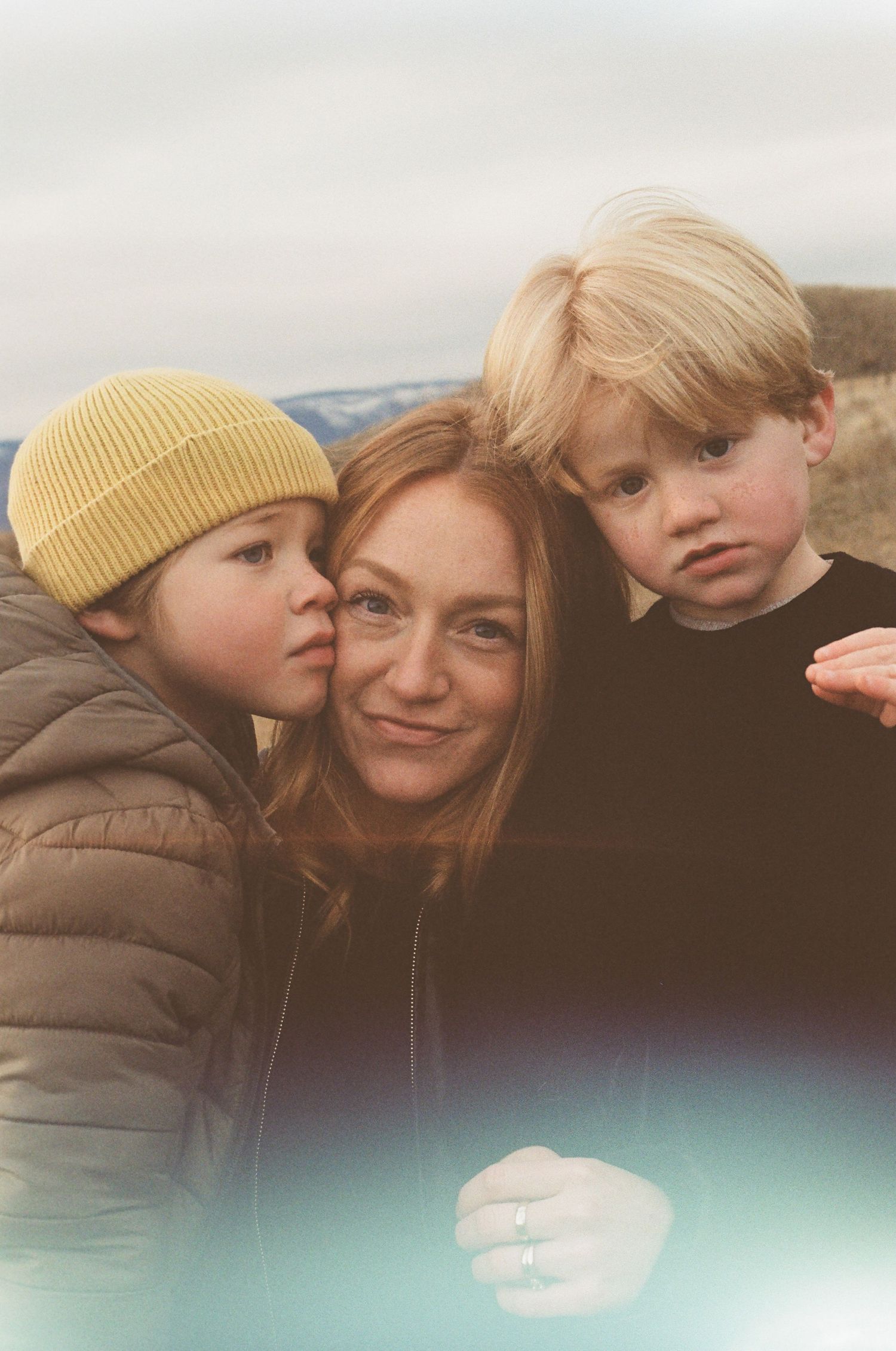 A mother smiles as two young blonde boys lean in for a cuddle outdoors on a cloudy day.
