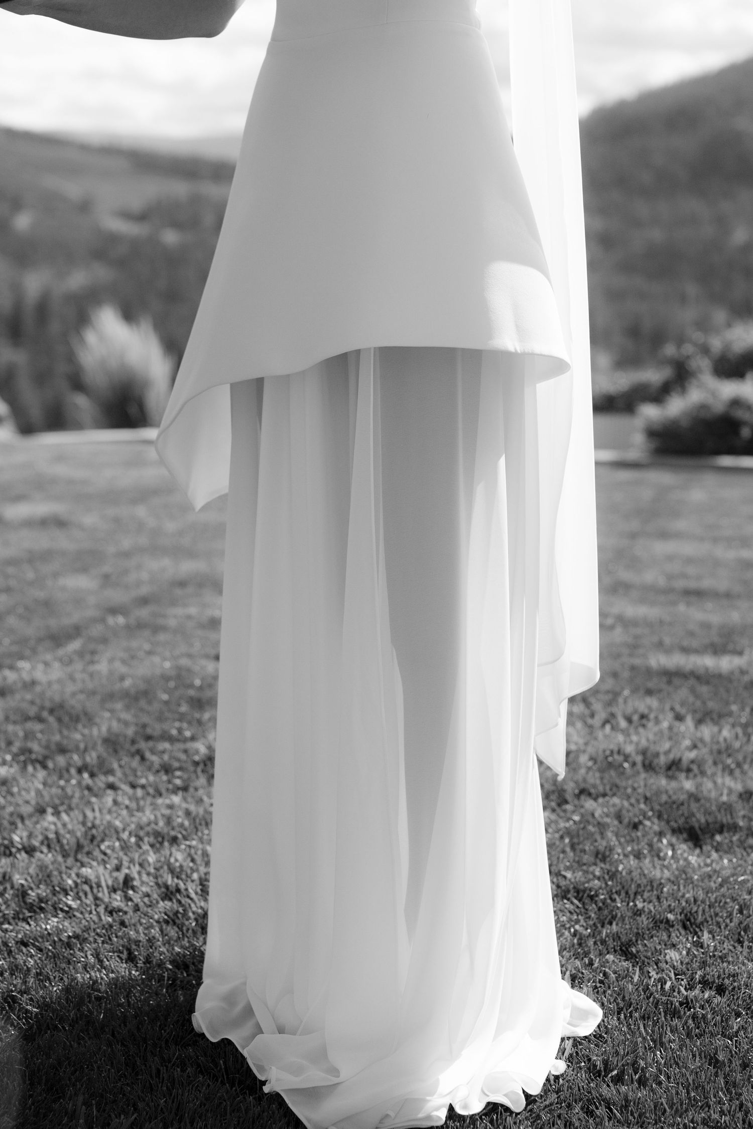 Close-up of a bride in a modern white wedding gown with layered peplum top and flowing chiffon skirt outdoors.