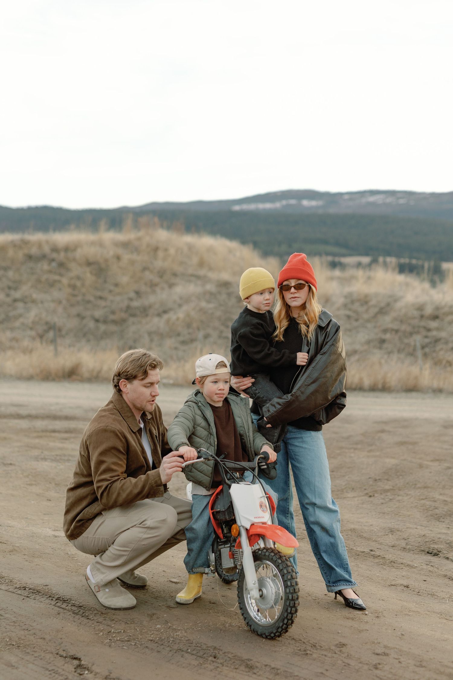 Family with two children gathered around a small dirt bike on a rural gravel road with hills in the background.