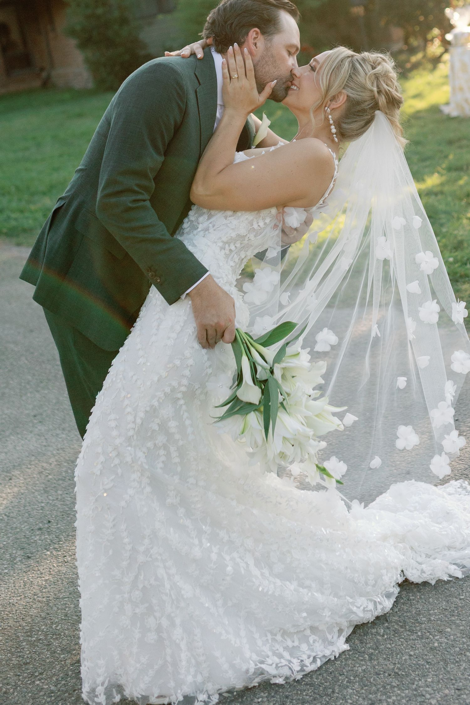 Bride and groom kiss romantically as floral veil drapes around them outdoors.