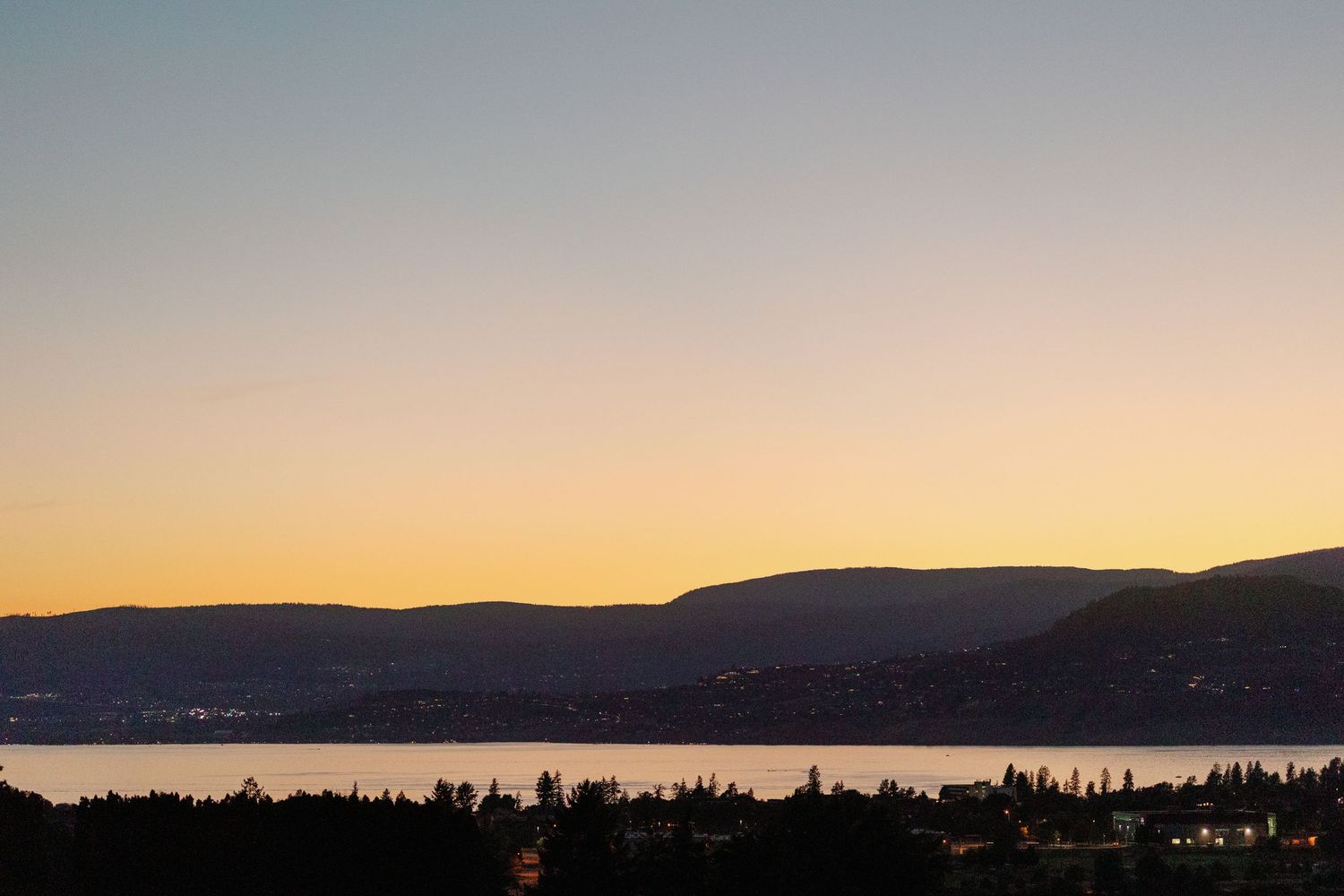 Golden sunset over calm lake with mountain silhouette and city lights below.