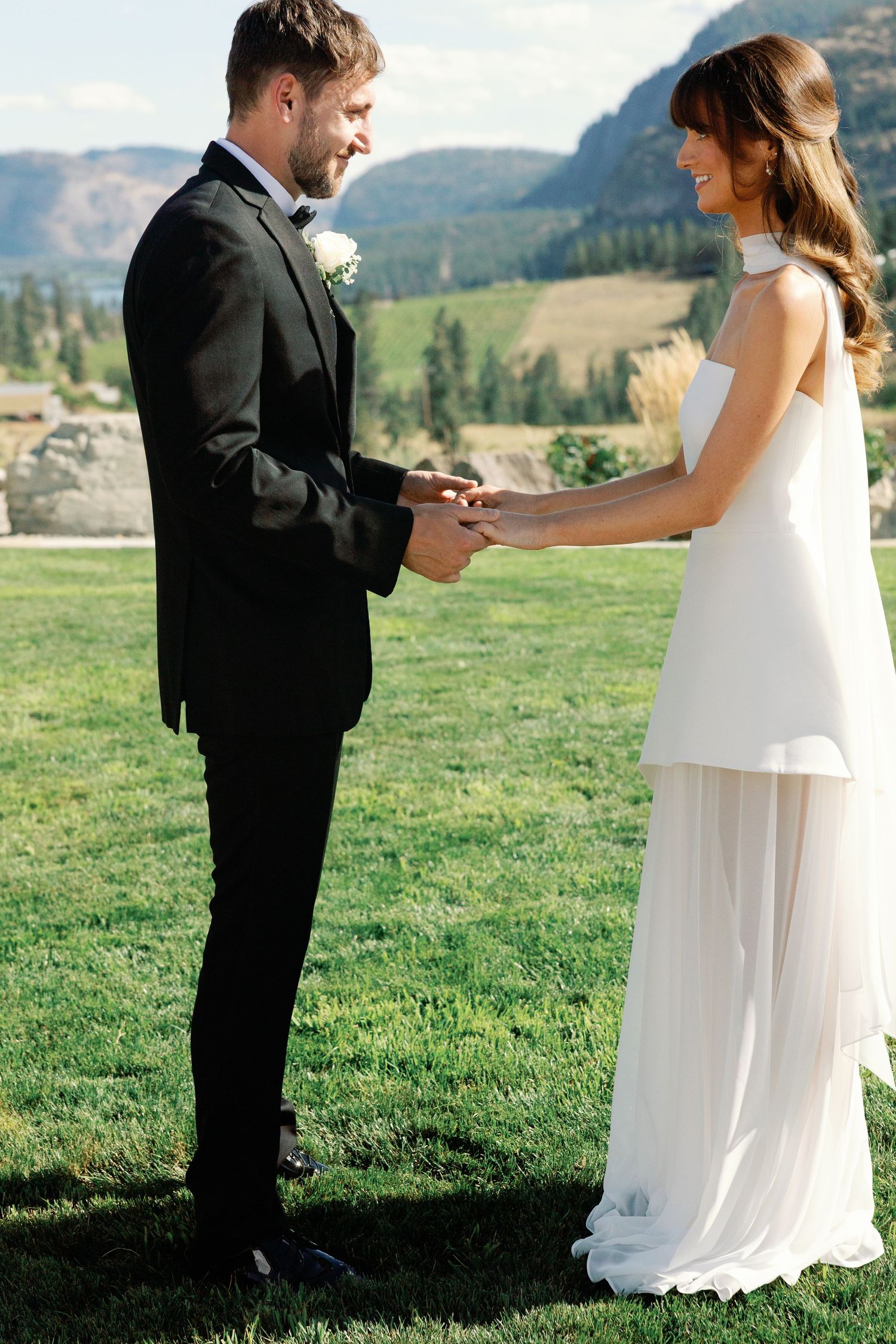 Bride and groom laugh while exchanging rings on green lawn with stunning mountain backdrop.