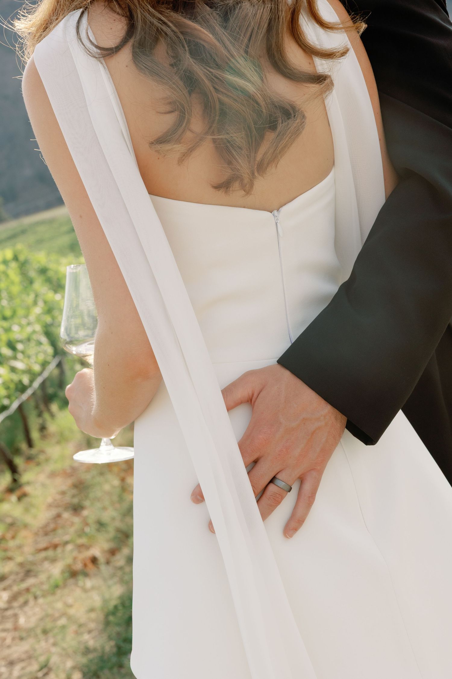 Groom in black suit embraces bride in white wedding dress from behind, showing wedding band near vineyard.